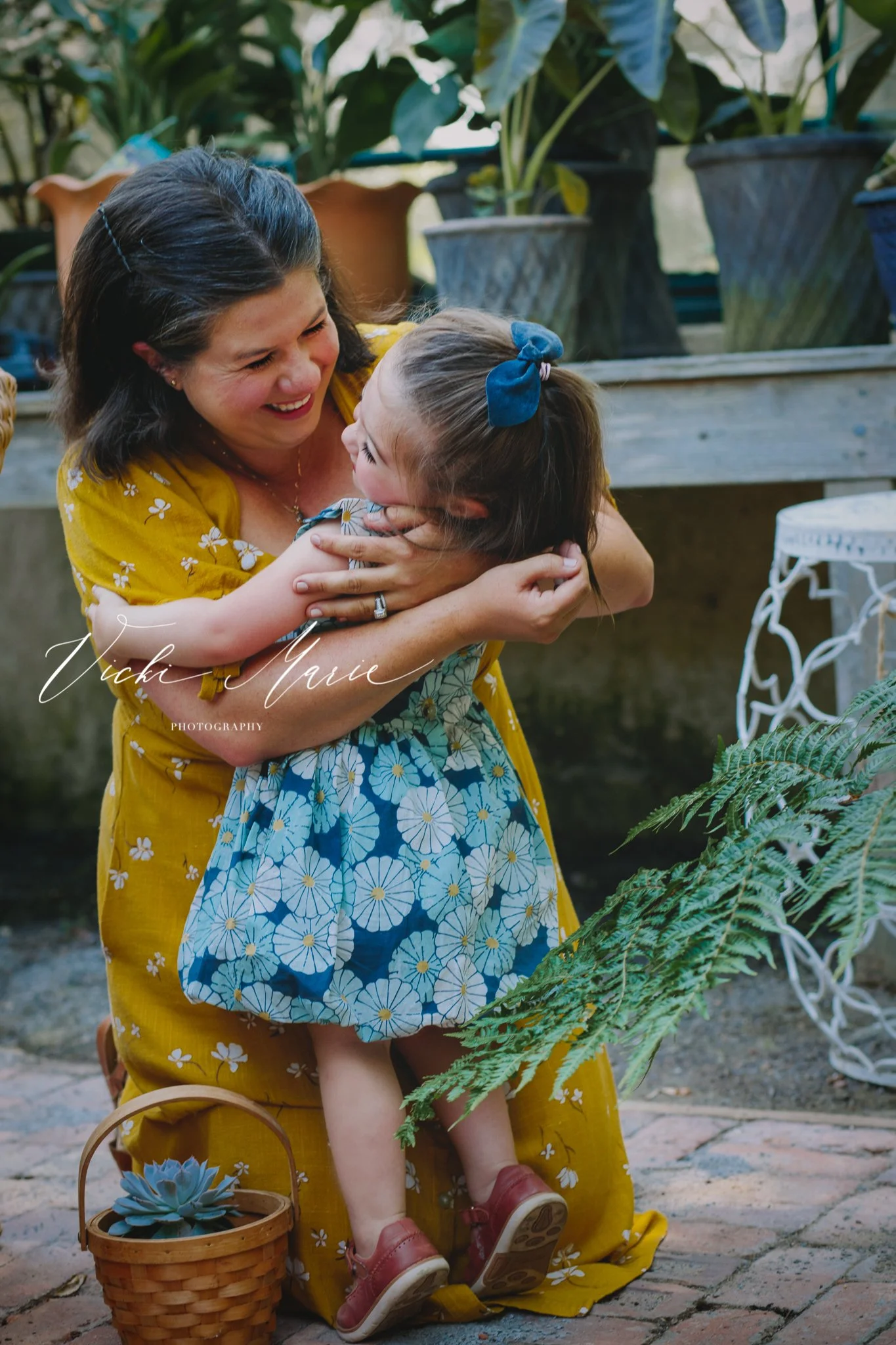 A woman in a yellow dress holding and hugging a young girl in a blue floral dress, both smiling and showing affection in an outdoor garden setting.