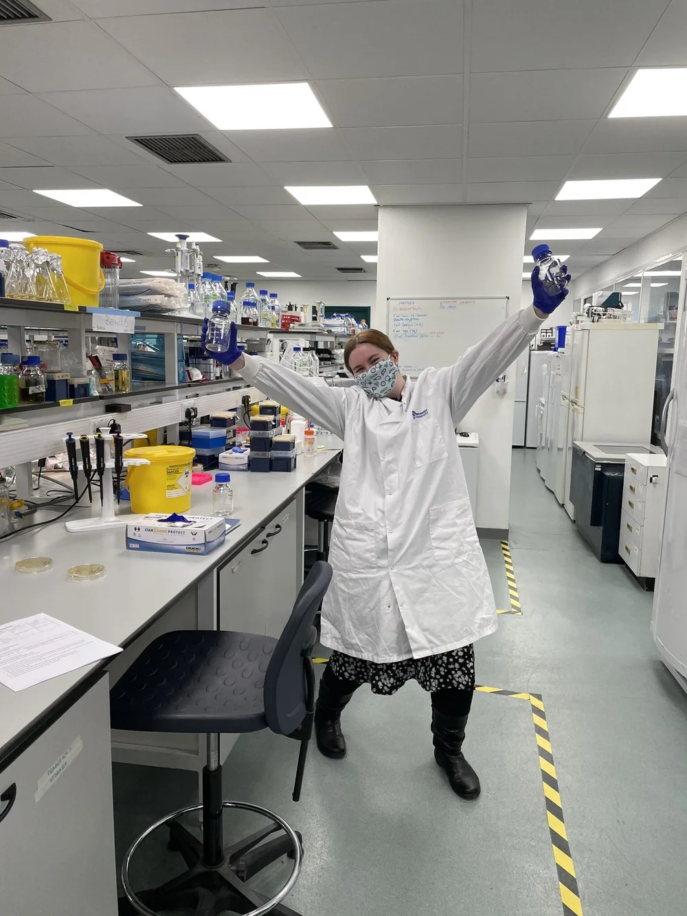  Me in my current lab where I am doing my PhD. The photo is a full-body shot of me enthusiastically posing with two Duran bottles. I am wearing a white lab coat and a pale blue mask. 