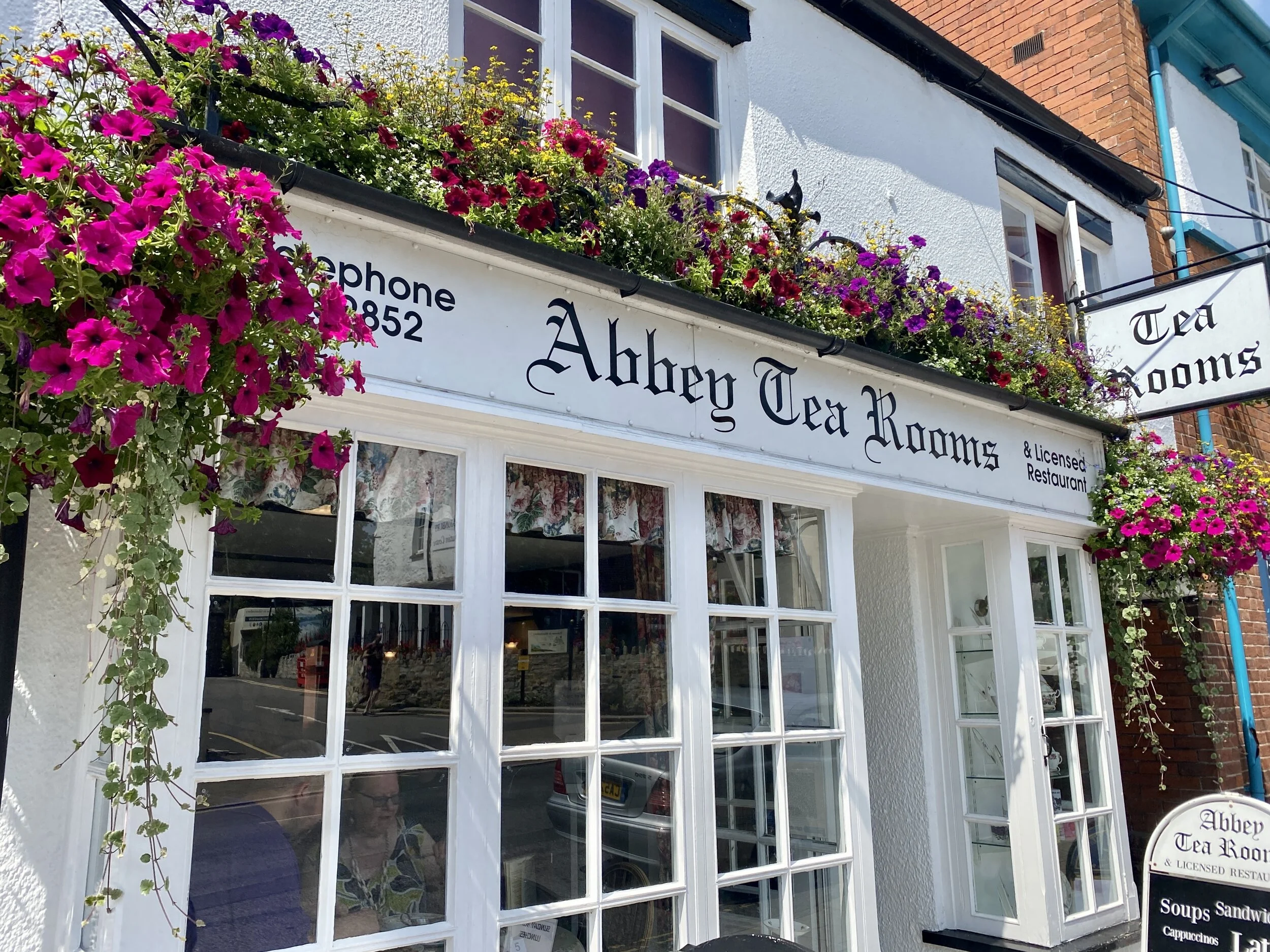 The storefront of Abbey Tea Rooms and Licensed Restaurant decorated with colorful flowers in window boxes, with signs displaying the restaurant's name and menu items on a sidewalk chalkboard outside.