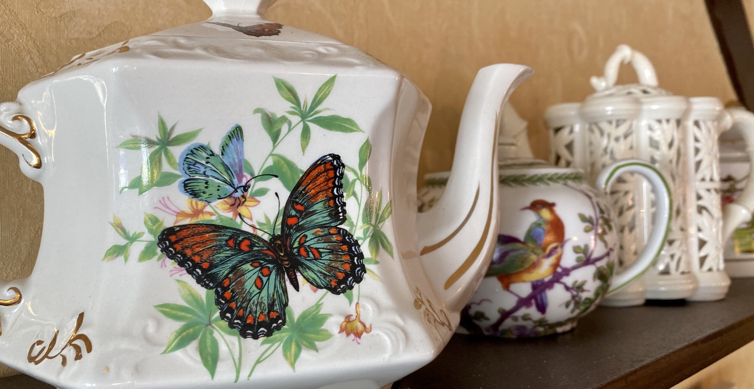 Close-up of a white porcelain teapot decorated with colorful butterflies and green leaves, with additional teapots and pitchers in the background.