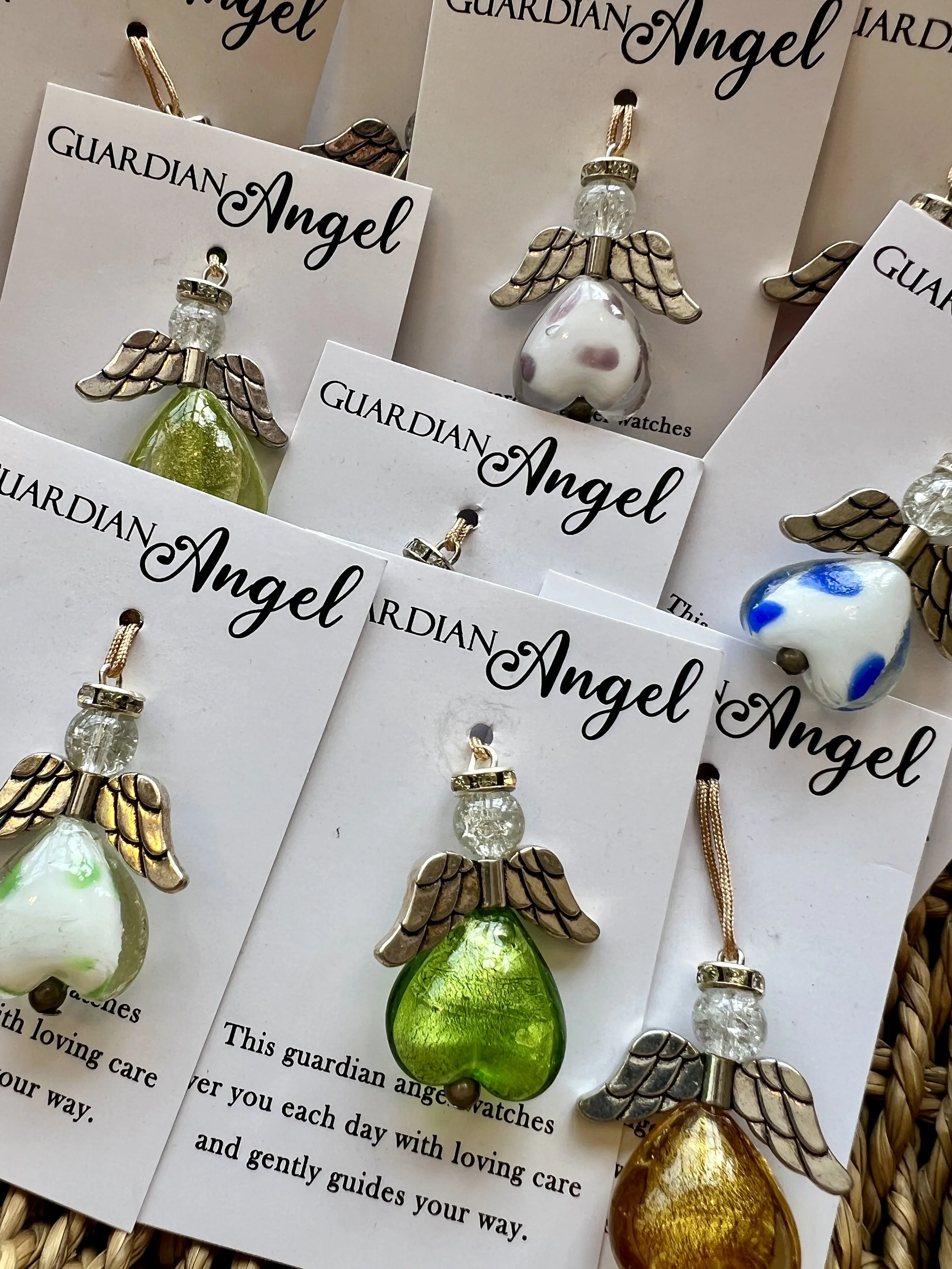 Multiple glass guardian angel figurines with wings and beads, attached to white cards with black cursive text that reads 'Guardian Angel.' The figurines are various colors including green, blue, yellow, and clear.