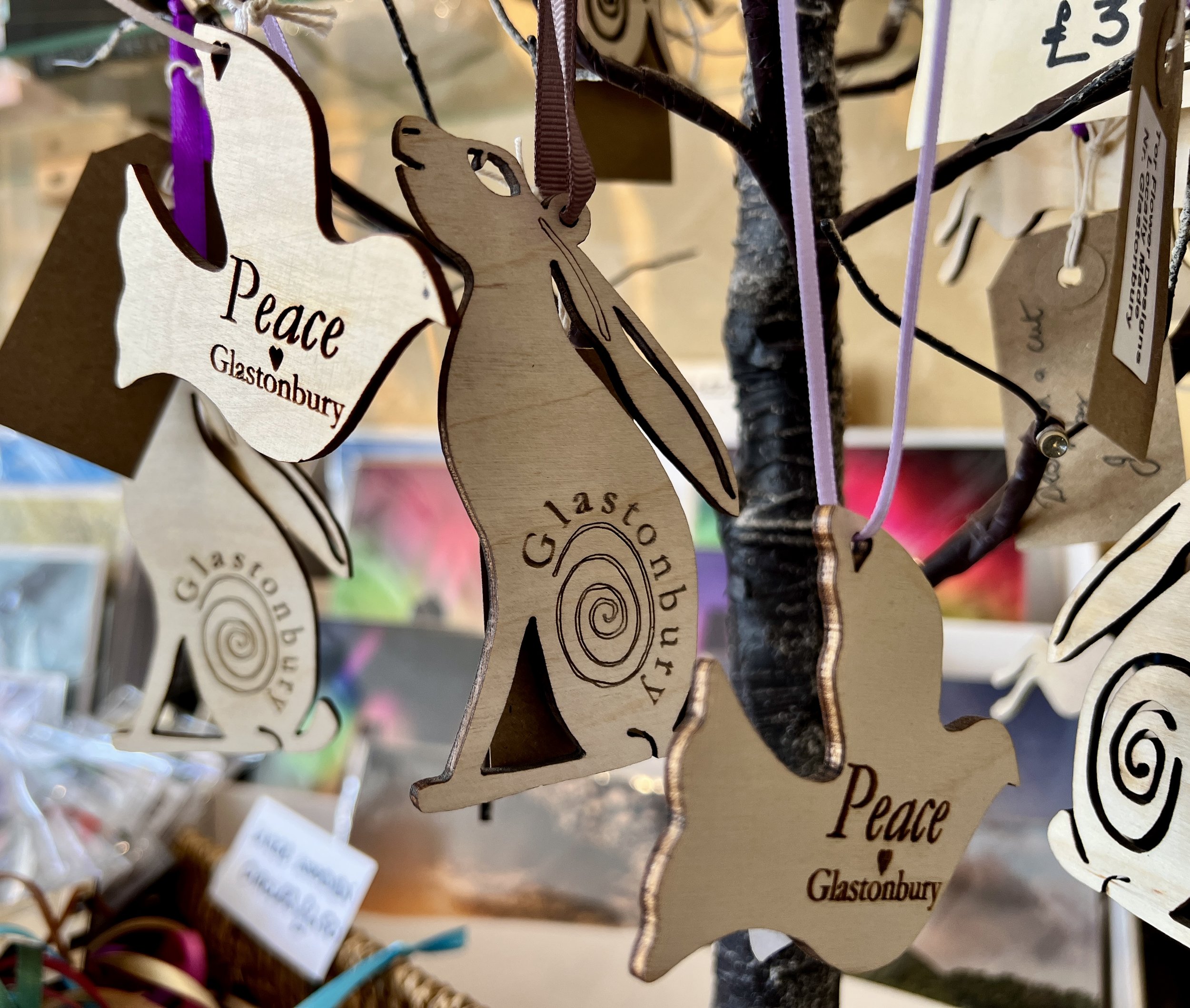 Wooden ornaments shaped like hearts and leaves hanging from a tree branch, with words like 'Peace' and 'Glastonbury' engraved on some pieces, decorated with ribbons in purple, lavender, and brown.
