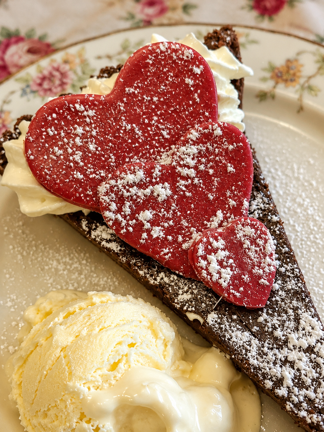 A slice of chocolate cake topped with white whipped cream, three red heart-shaped cookies dusted with powdered sugar, and a scoop of vanilla ice cream on a floral plate.