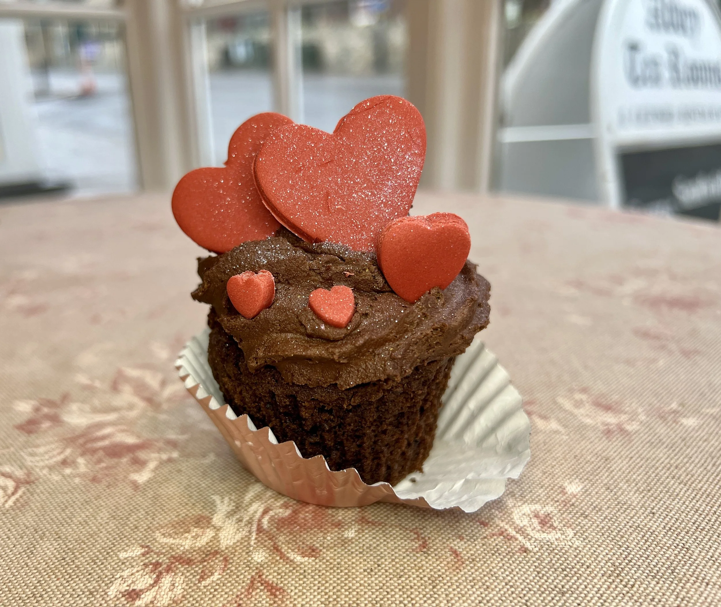 A cupcake topped with red heart-shaped decorations and a glittery red heart on top, sitting on a beige tablecloth.