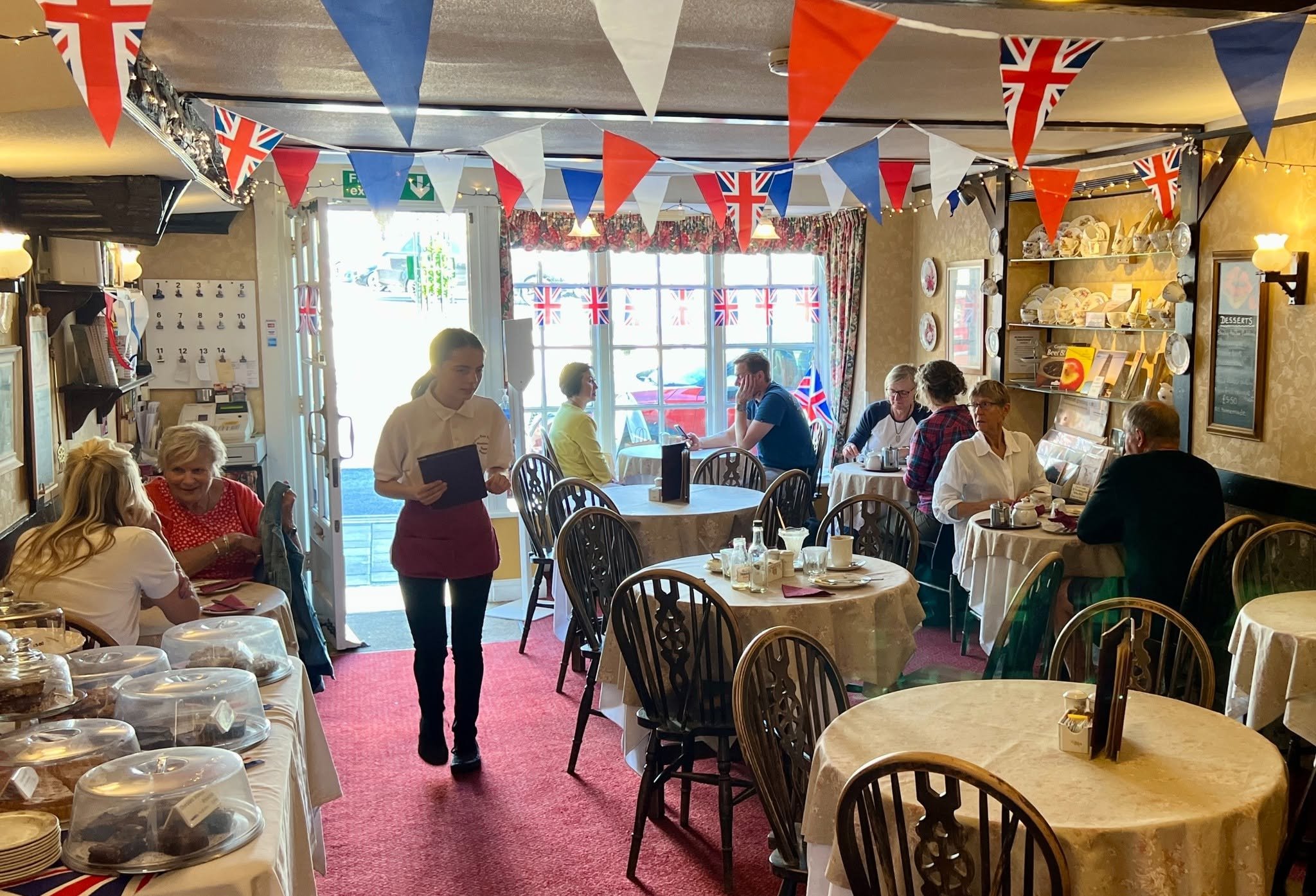 A lively restaurant decorated for a celebration with British flags and bunting hanging from the ceiling, filled with patrons dining and a waitress serving tables.