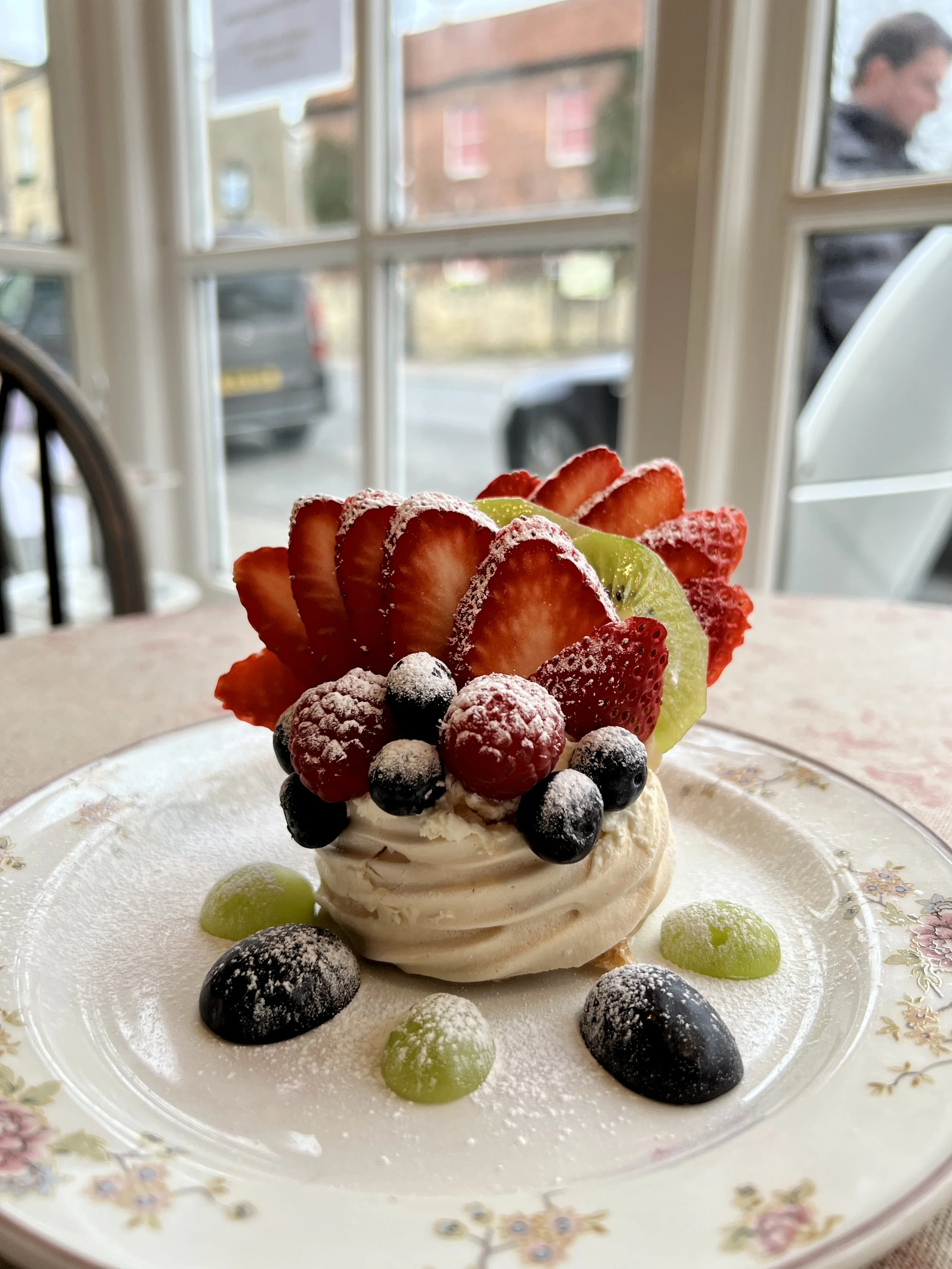 A dessert with various sliced strawberries, blueberries, green grapes, and a dollop of whipped cream or frosting on a decorative floral plate, with a window and outdoor scene in the background.