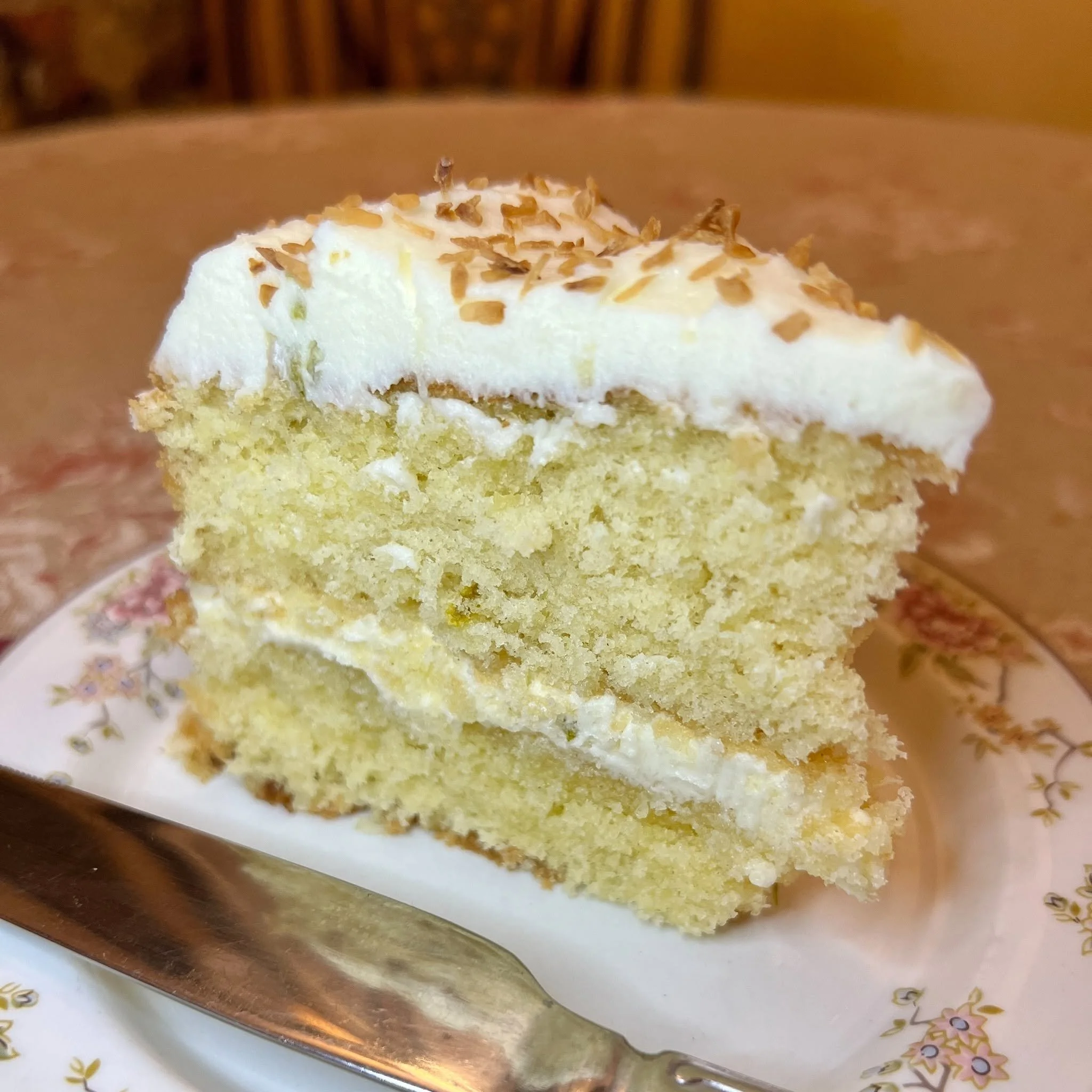 A slice of cake with white frosting and chopped nuts on top, on a floral-patterned plate with a silver fork.
