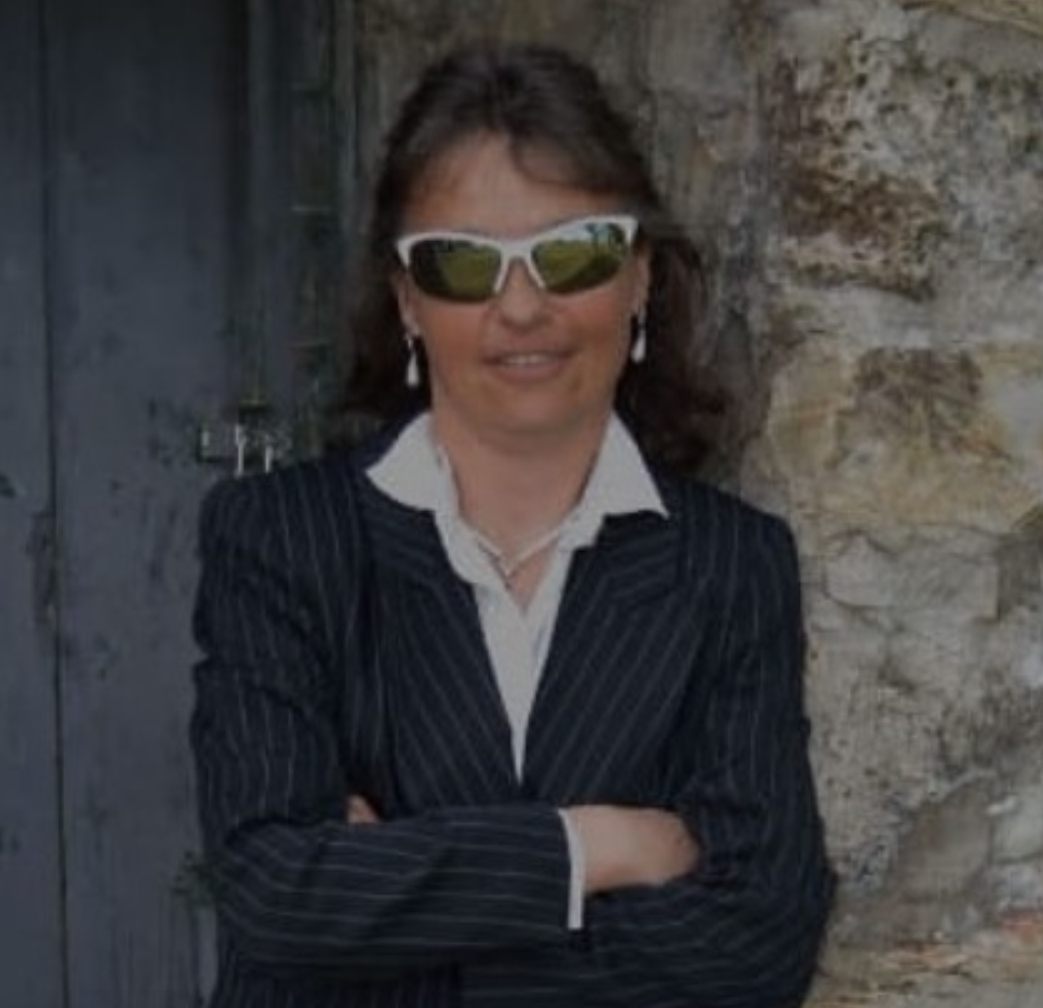 A woman with shoulder-length brown hair wearing sunglasses, earrings, a white shirt, a dark pinstripe blazer, and standing with arms crossed in front of a rustic stone wall.