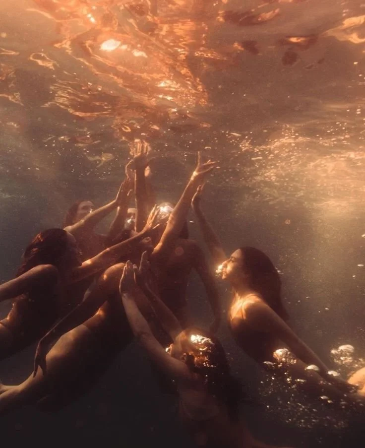 Group of women underwater, reaching upward and touching each other's hands, illuminated by warm light.