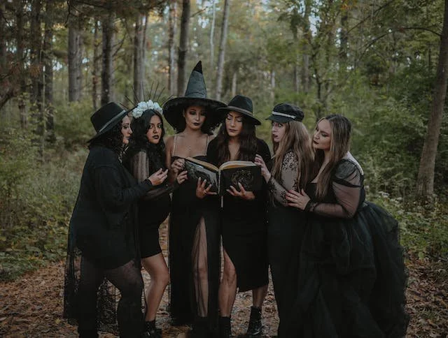 A group of witches wearing black all looking at a book while in the forest