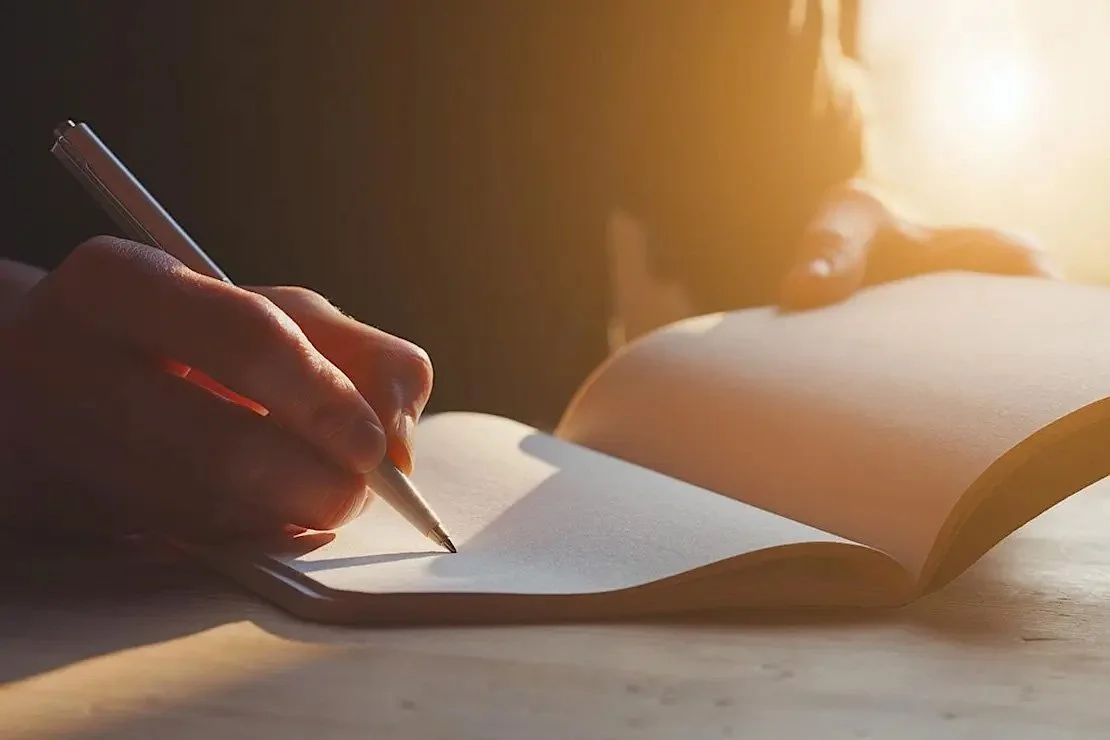 close up of a person using a pen in a notebook with a warm cast of sunlight over the photo