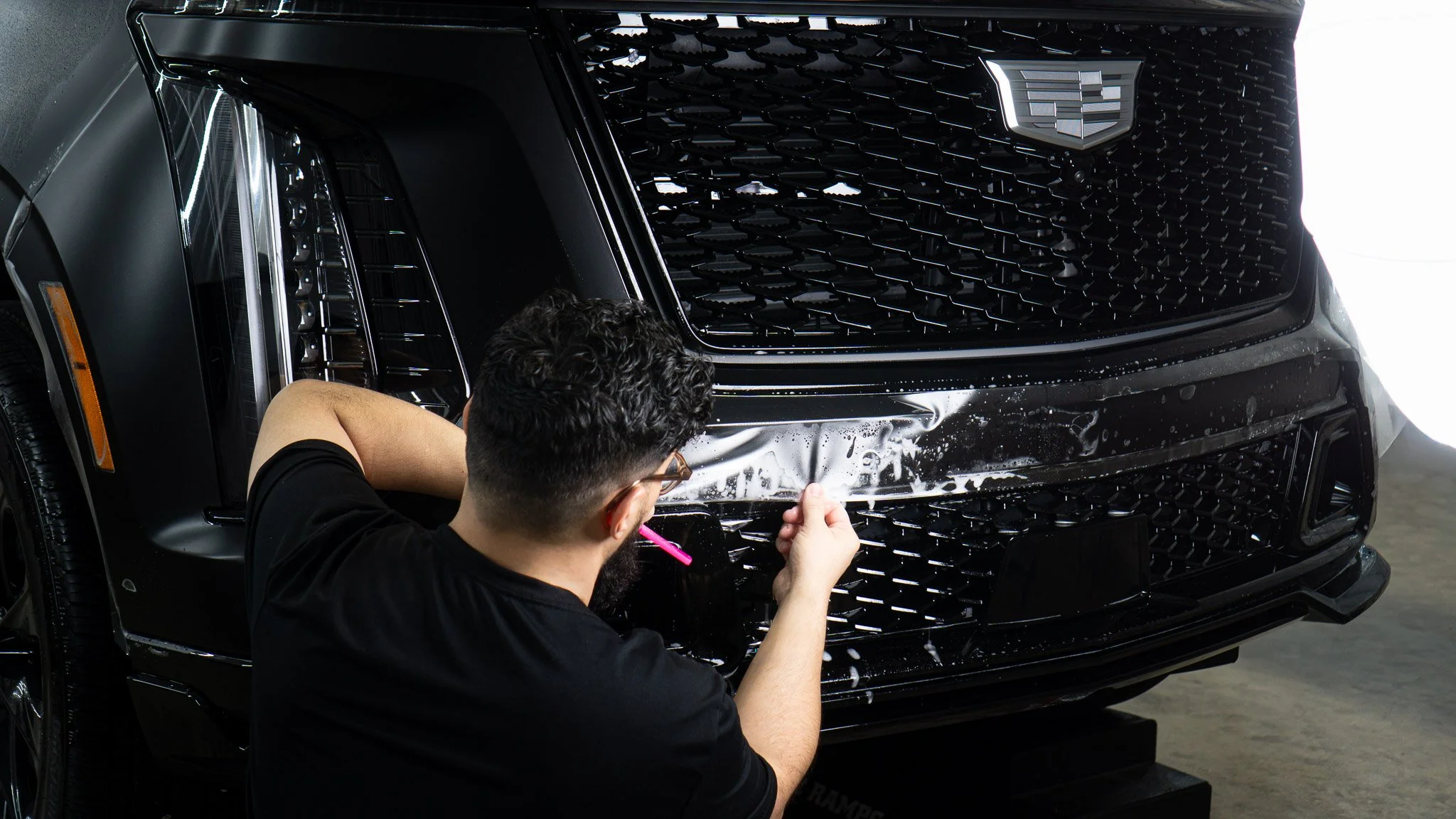 Technician applying clear paint protection film to the front bumper of a black Cadillac Escalade at EIGHTYNINE VWS, Little Elm, TX — STEK-USA certified PPF installation in North Texas.