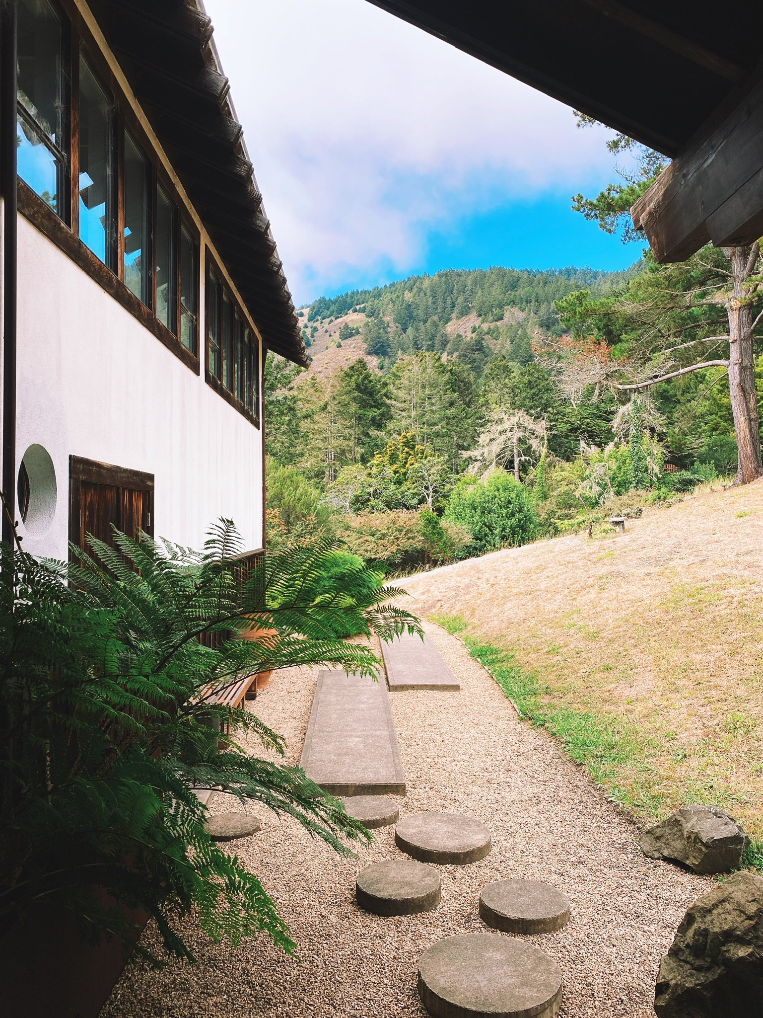 Kenko Retreat - a traditional Japanese-style building with a garden pathway that includes round stepping stones and benches, surrounded by lush trees and coastal mountains.