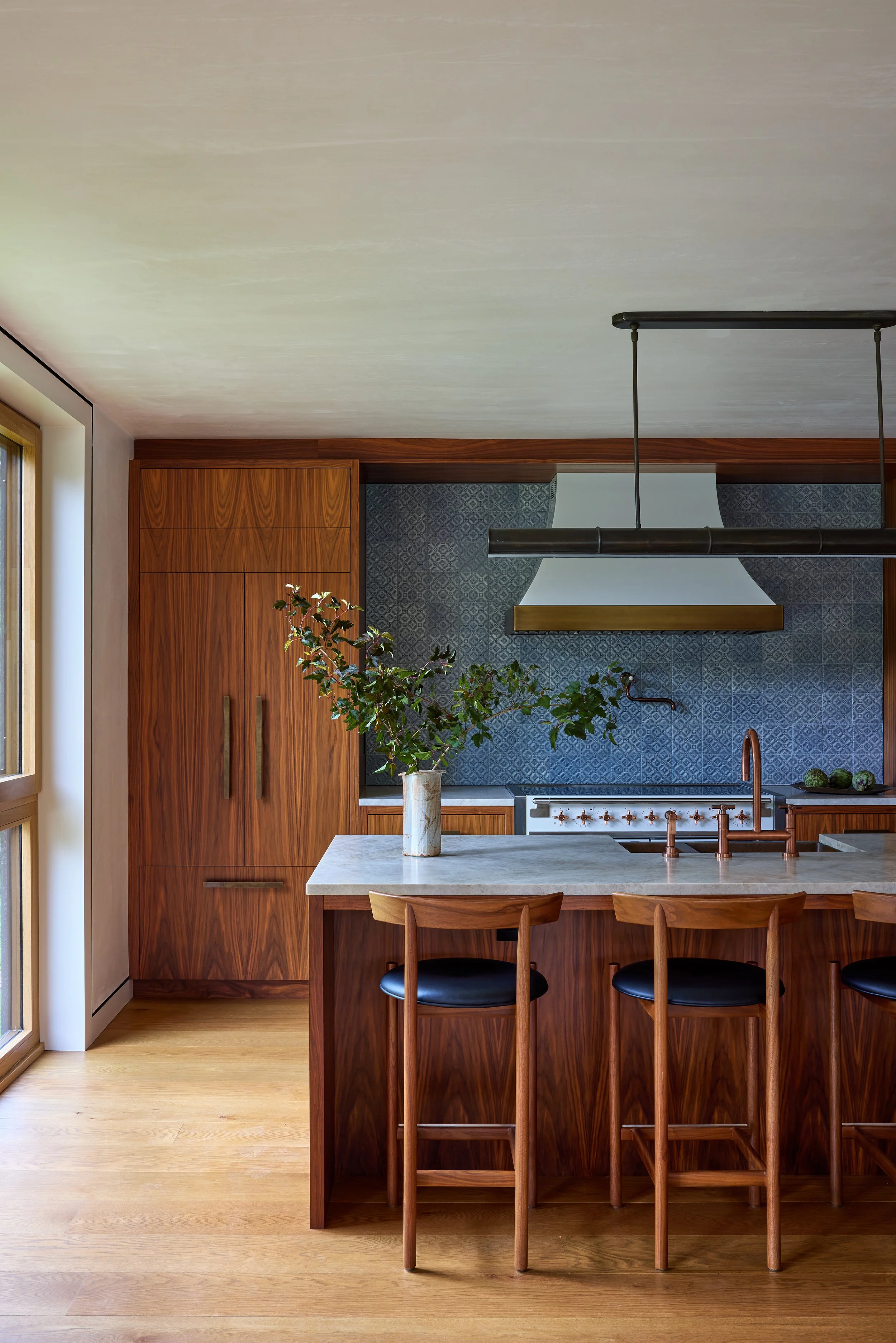 Modern kitchen with wooden cabinets, a white countertop, a large vase with green branches, and a breakfast bar with three wooden chairs.