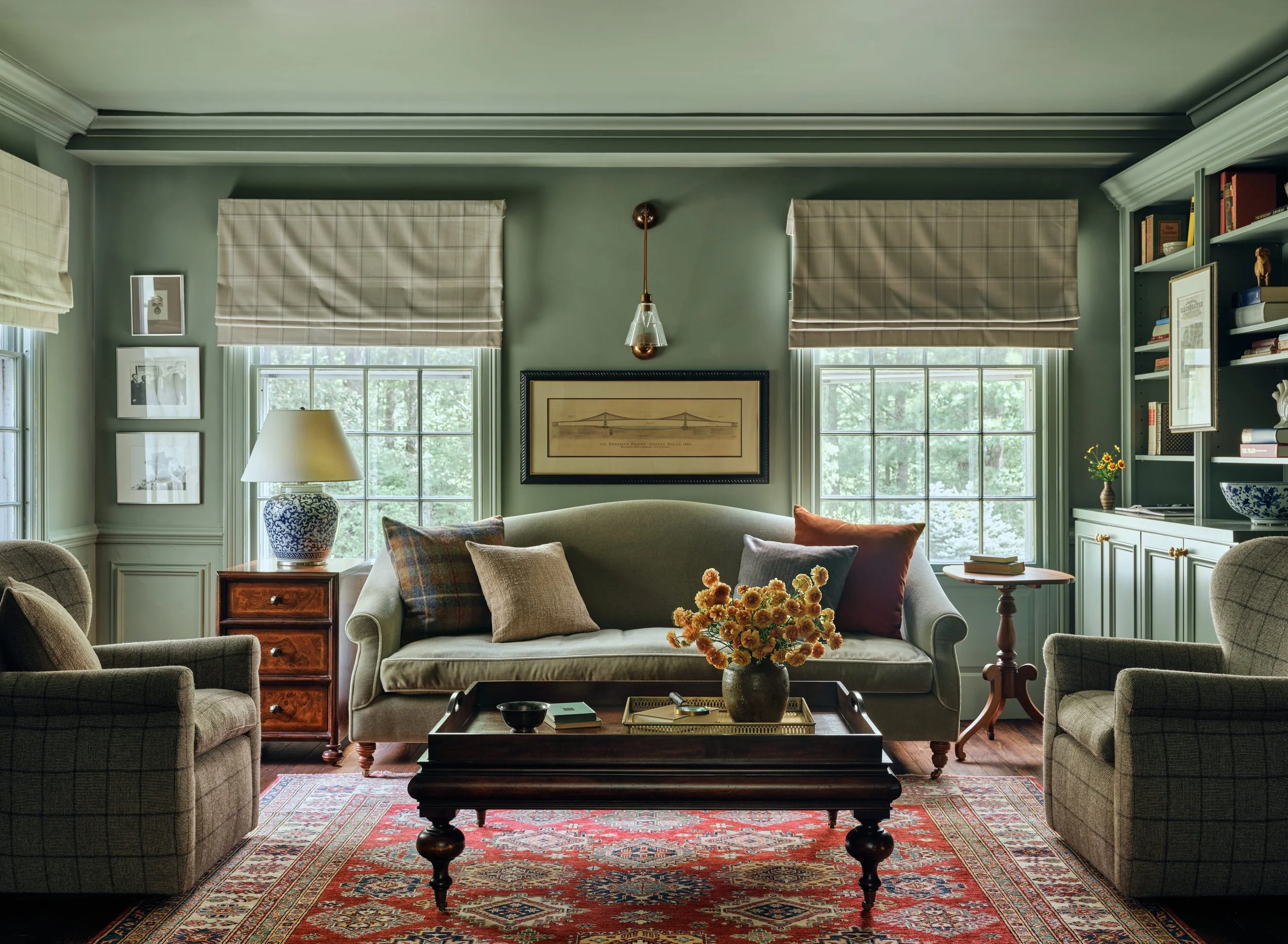 Living room with green walls, beige sofa with multiple pillows, armchairs, wooden coffee table with vase of flowers, bookshelf, and windows with beige blinds.