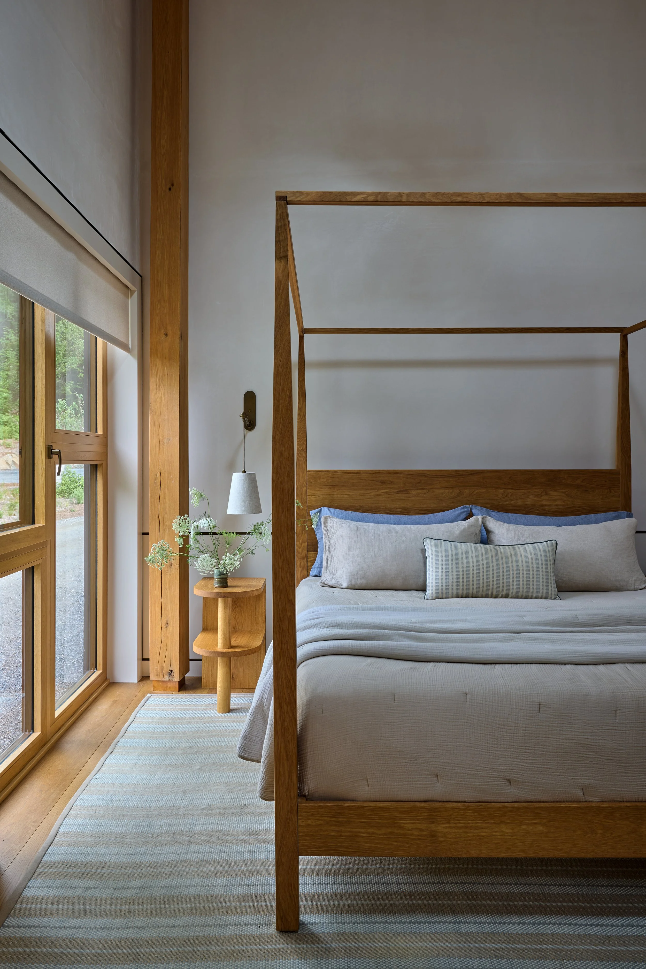 A bedroom with a wooden bed frame, white bedding, and a striped throw pillow. There is a small wooden side table with a vase of flowers and a wall-mounted light fixture. Large windows with wooden frames and a view of greenery outside.
