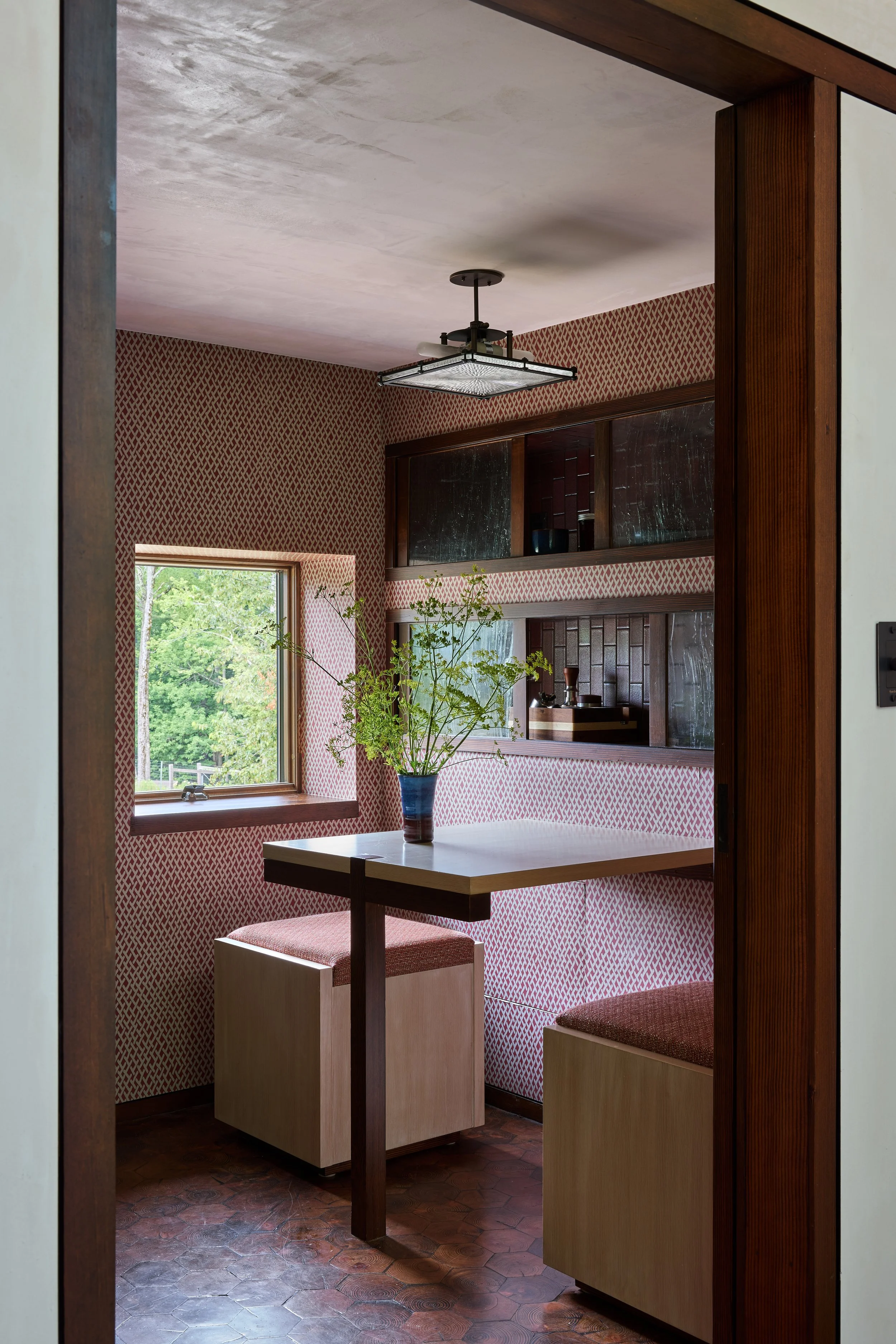 Cozy dining nook with pink patterned wallpaper, a small window showing green trees outside, a white table with a vase of green foliage, and two upholstered cube seats.