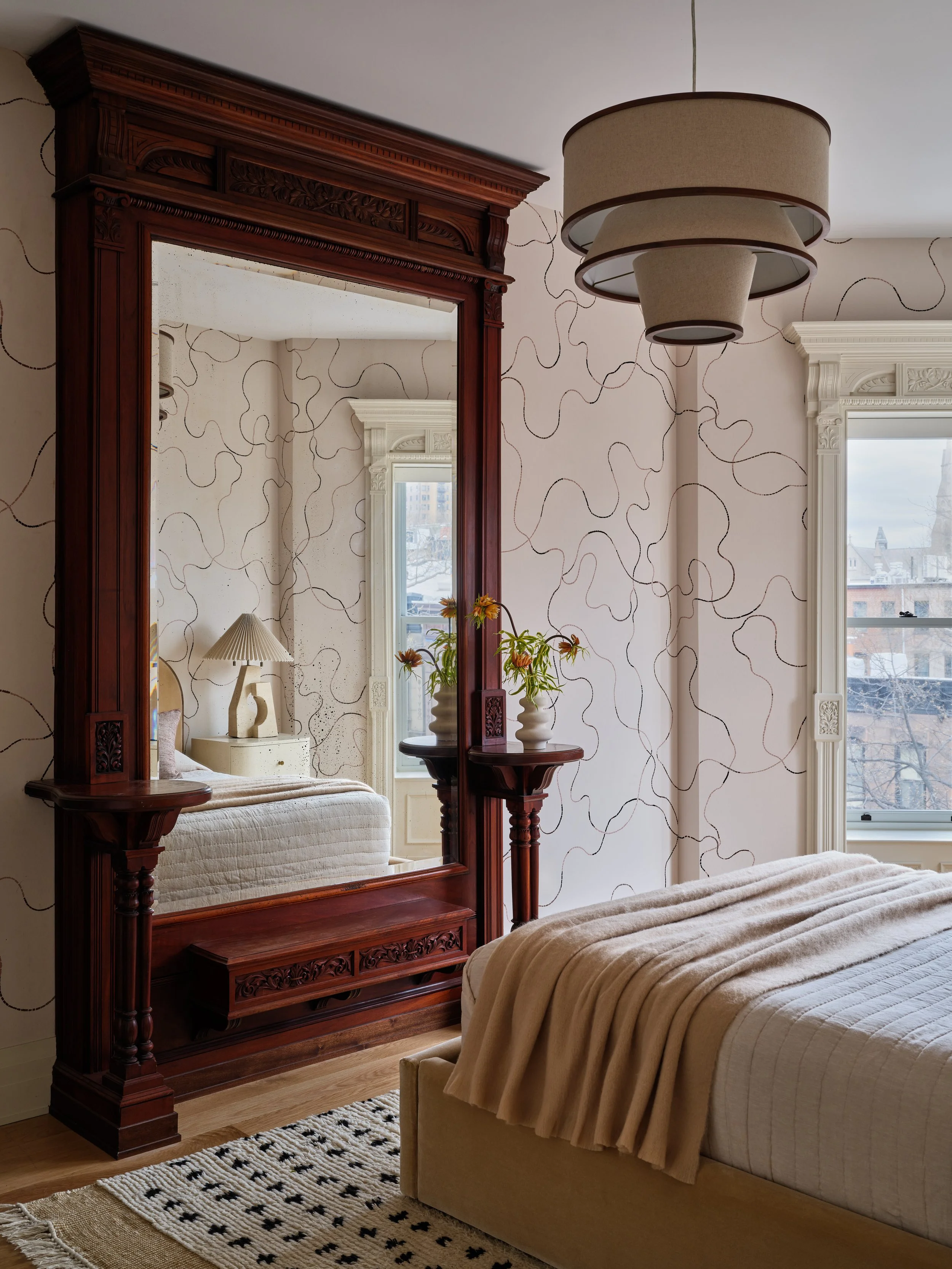 A bedroom with a large wooden mirror, a bed with cream bedding, a beige blanket, a patterned Rug, a window, and a modern pendant light fixture.