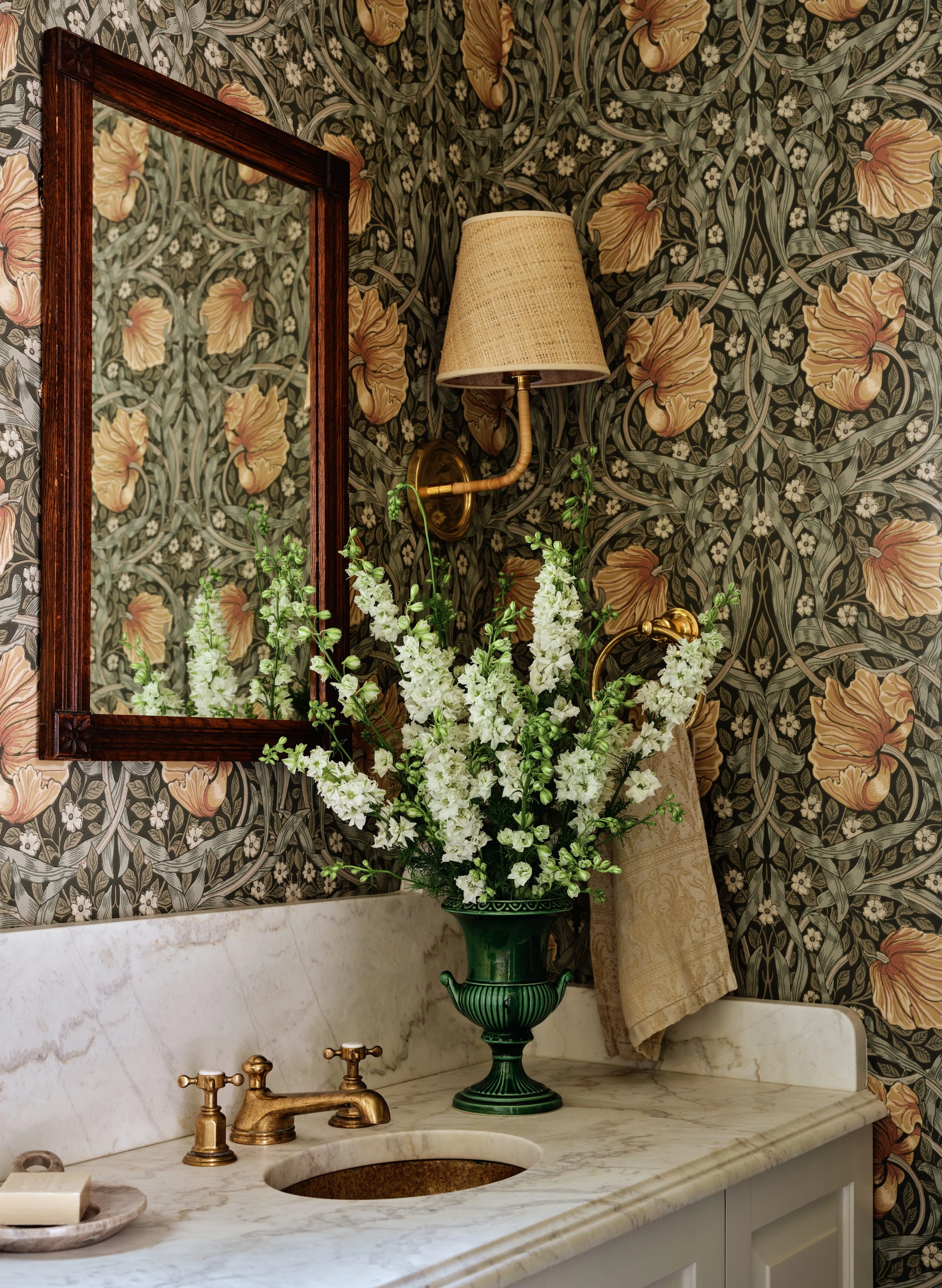 A bathroom vanity with a marble countertop, gold faucet, green vase with white flowers, a beige towel, a wall-mounted lamp with a beige shade, a mirror with a wooden frame, and floral wallpaper with large peach and green patterns.