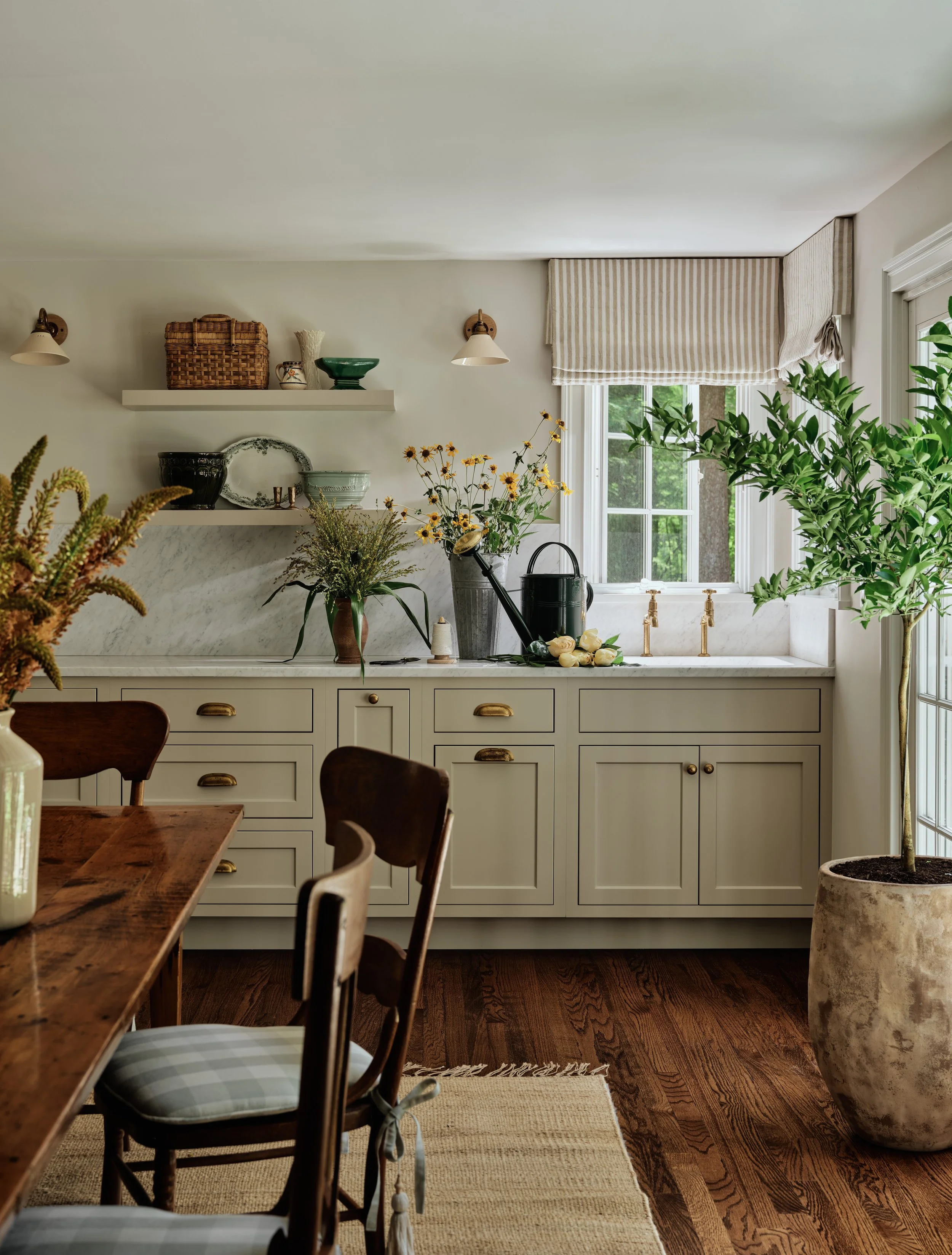 A cozy kitchen with white cabinets, a marble backsplash, and a window with striped blinds. The countertop holds potted plants, flowers, and gardening tools. Part of a wooden dining table with cushioned chairs is visible in the foreground.