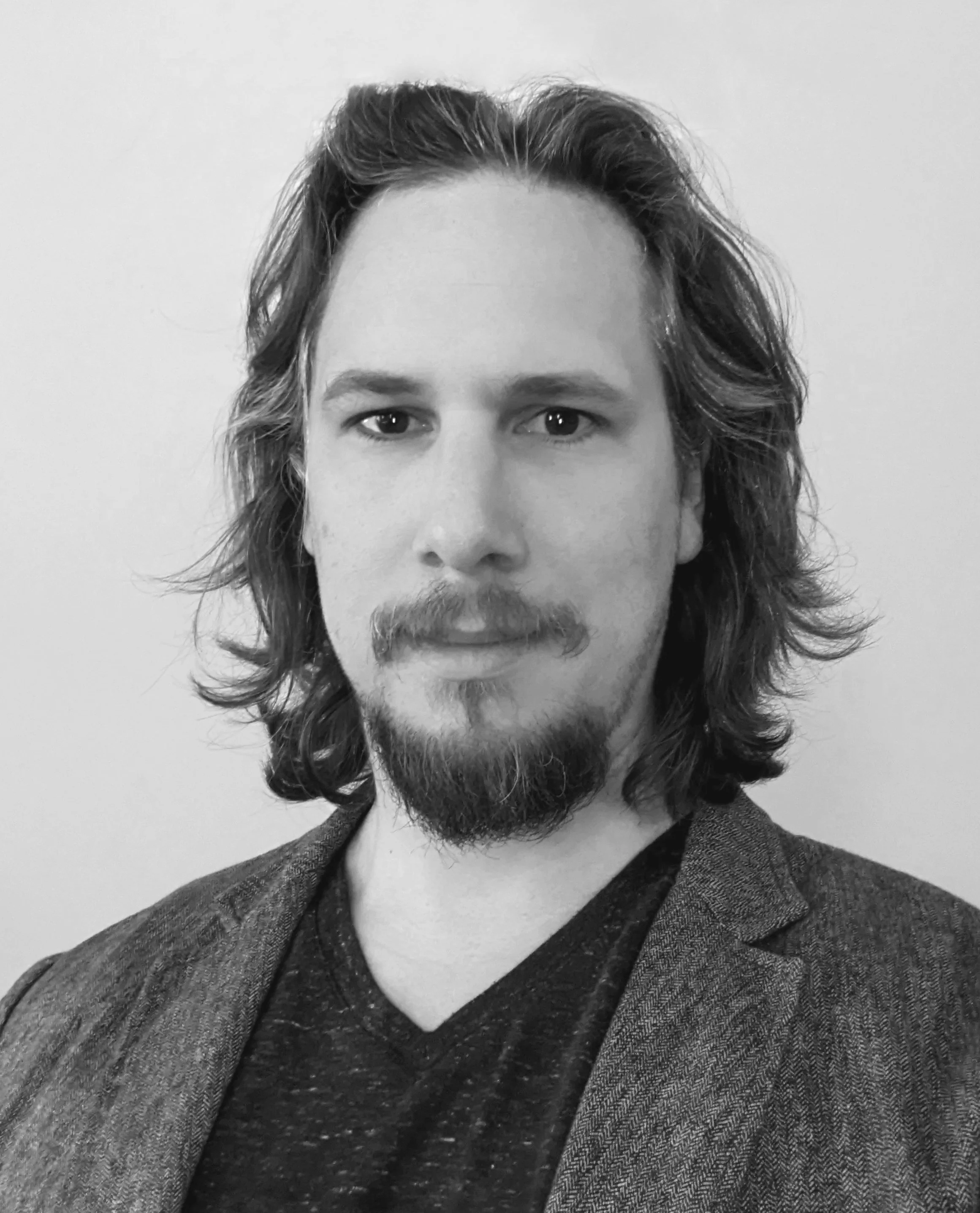 Black and white photograph of a young man with shoulder-length wavy hair and a beard, wearing a dark v-neck shirt and a textured blazer, against a plain background.