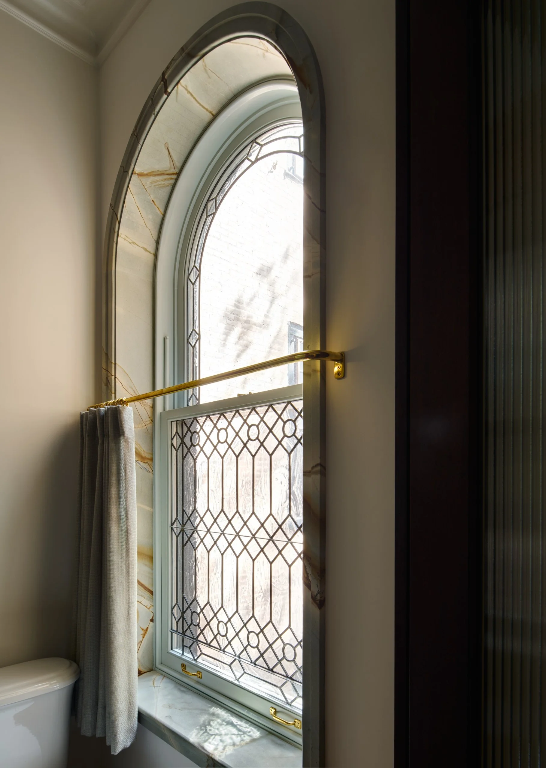 A bathroom window with decorative glass and brass handles, a marble frame, and beige curtains, with part of a toilet visible below the window.