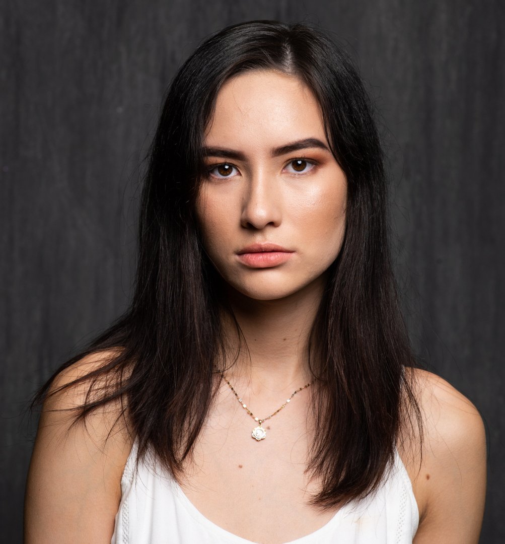 A young woman with long dark hair wearing a white top and a gold necklace with a pendant, standing against a dark background.