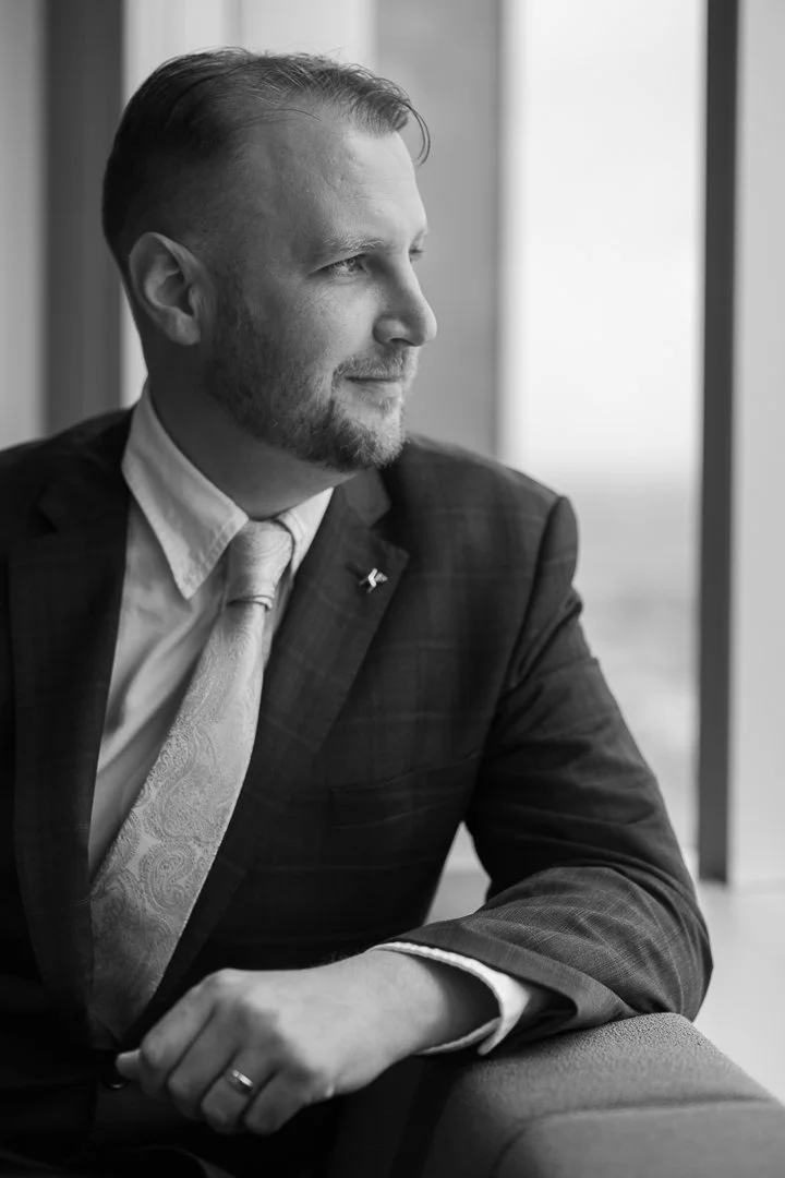 Man in formal business attire sitting by a window, looking outside, in black and white.