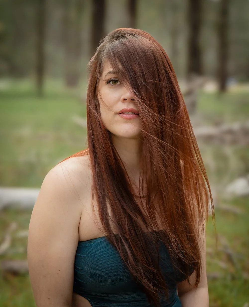 Portrait of a woman with long red hair partially covering her face, wearing a strapless blue top, outdoors with a blurred natural background.