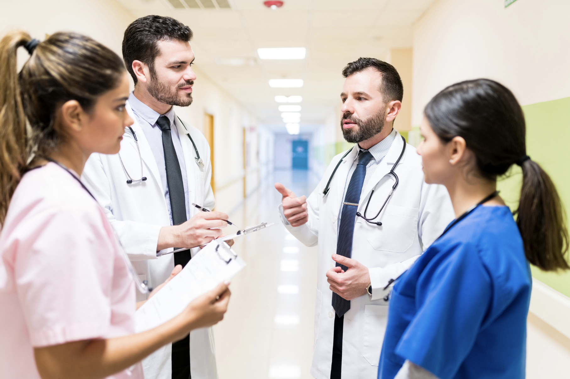 Group of four medical professionals, two men and two women, engaged in a discussion in a hospital corridor, with medical attire and stethoscopes.