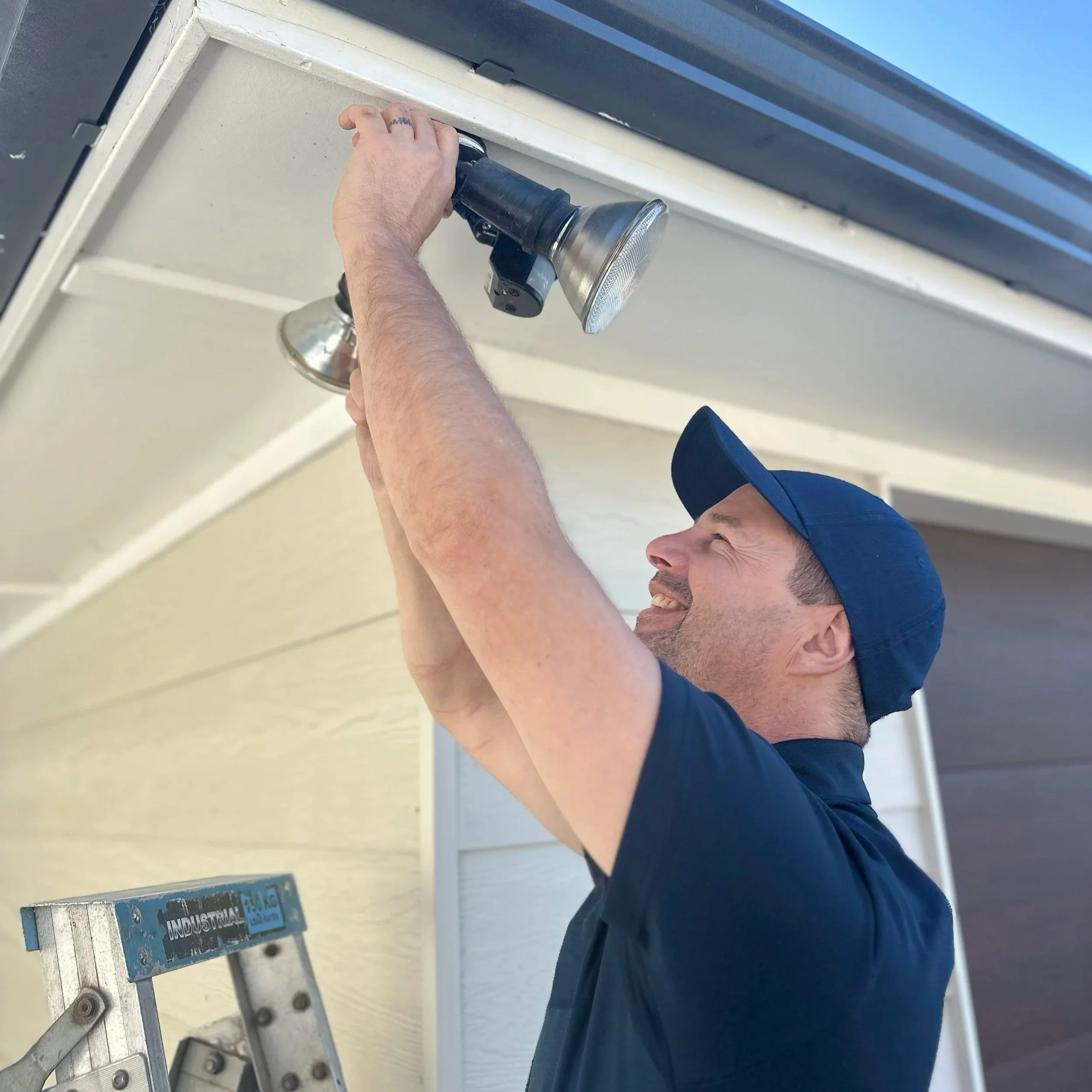 A man wearing a blue cap and navy shirt is installing or adjusting an outdoor light fixture under the eaves of a house.