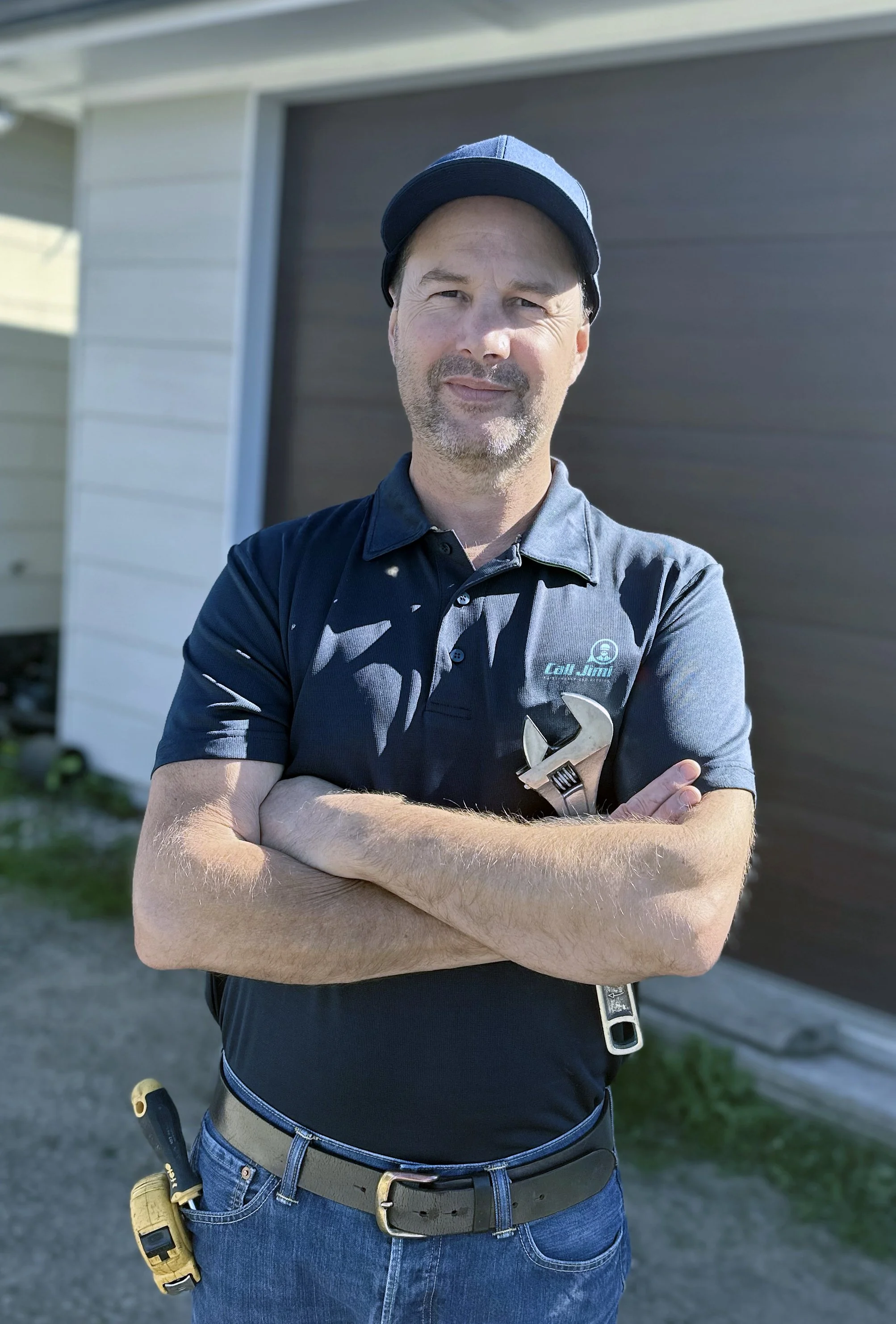 A man standing outdoors in front of a house with his arms crossed, wearing a navy blue cap, dark polo shirt with a logo, and holding a wrench, with a yellow screwdriver in his pocket.