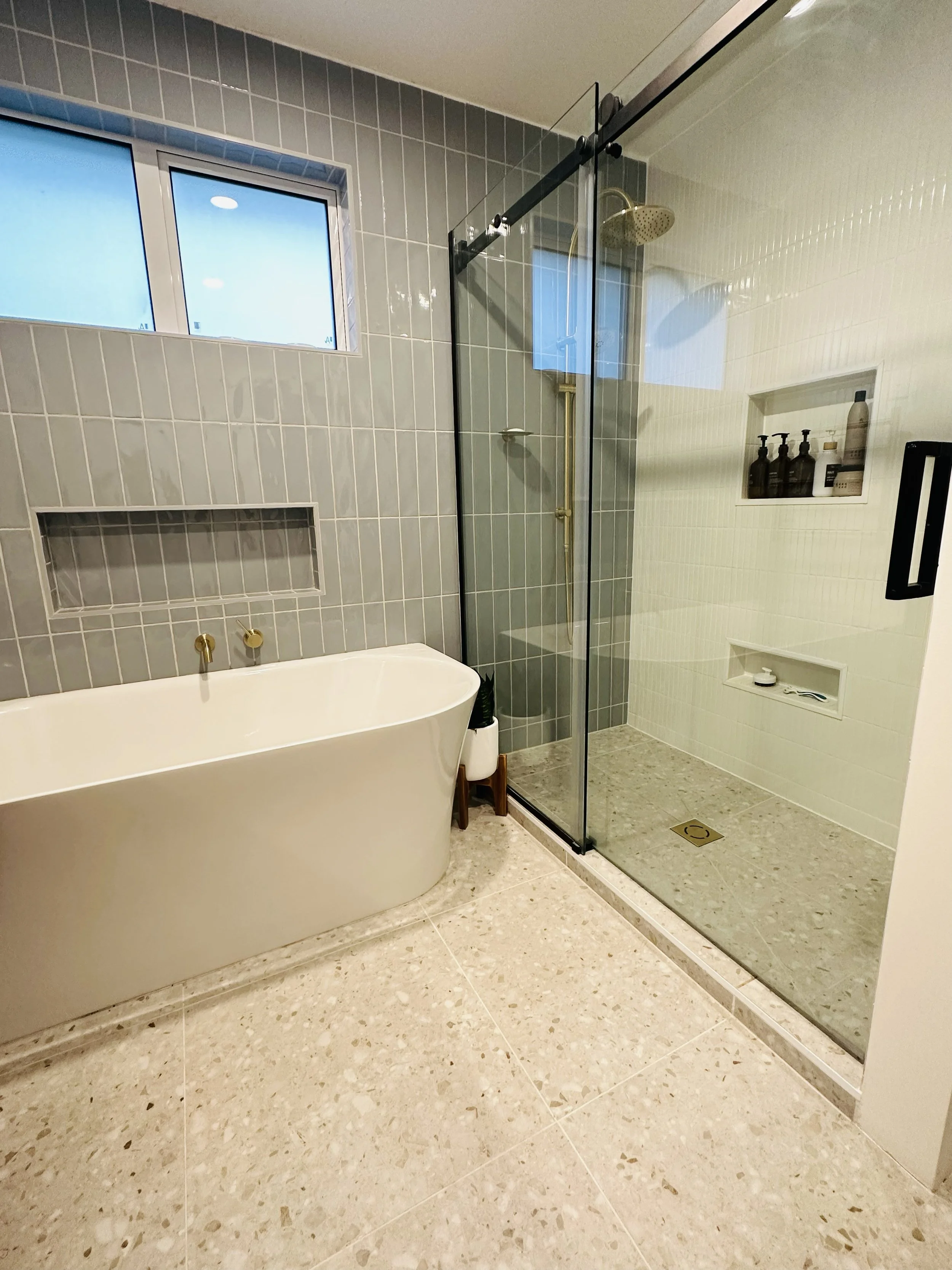 Modern bathroom with a white freestanding bathtub, a glass-enclosed shower with a brass showerhead, a window, and a built-in wall niche with toiletries.
