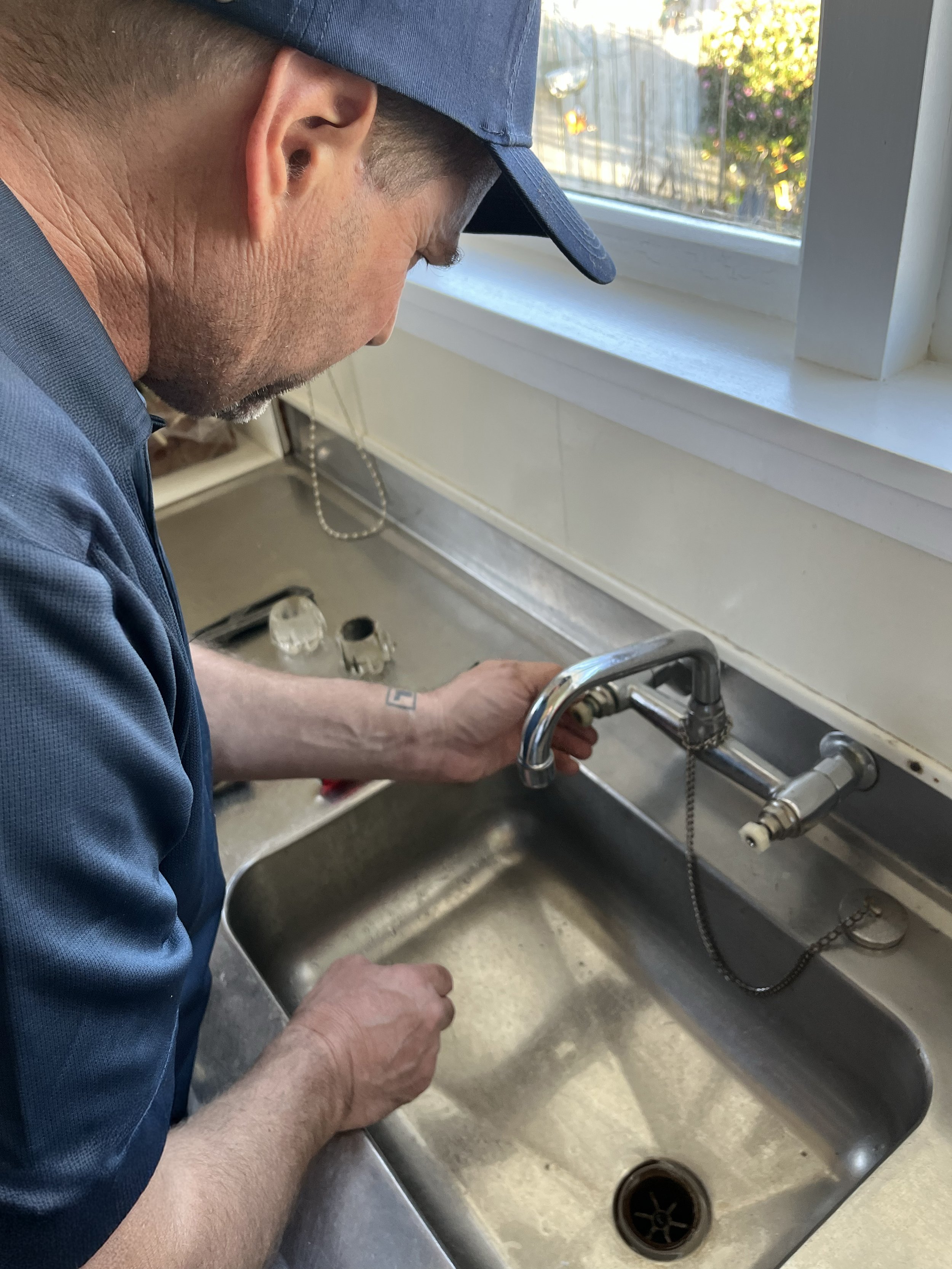 A man in a blue cap and uniform cleaning a stainless steel kitchen sink, with a window showing trees and sunlight in the background.