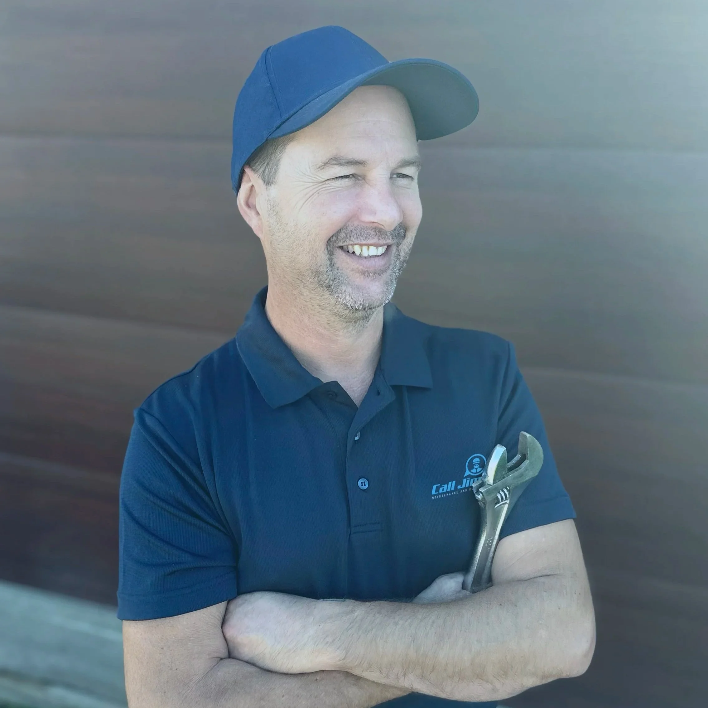 A man in a blue cap and polo shirt holding a wrench, smiling, with arms crossed, standing outdoors.