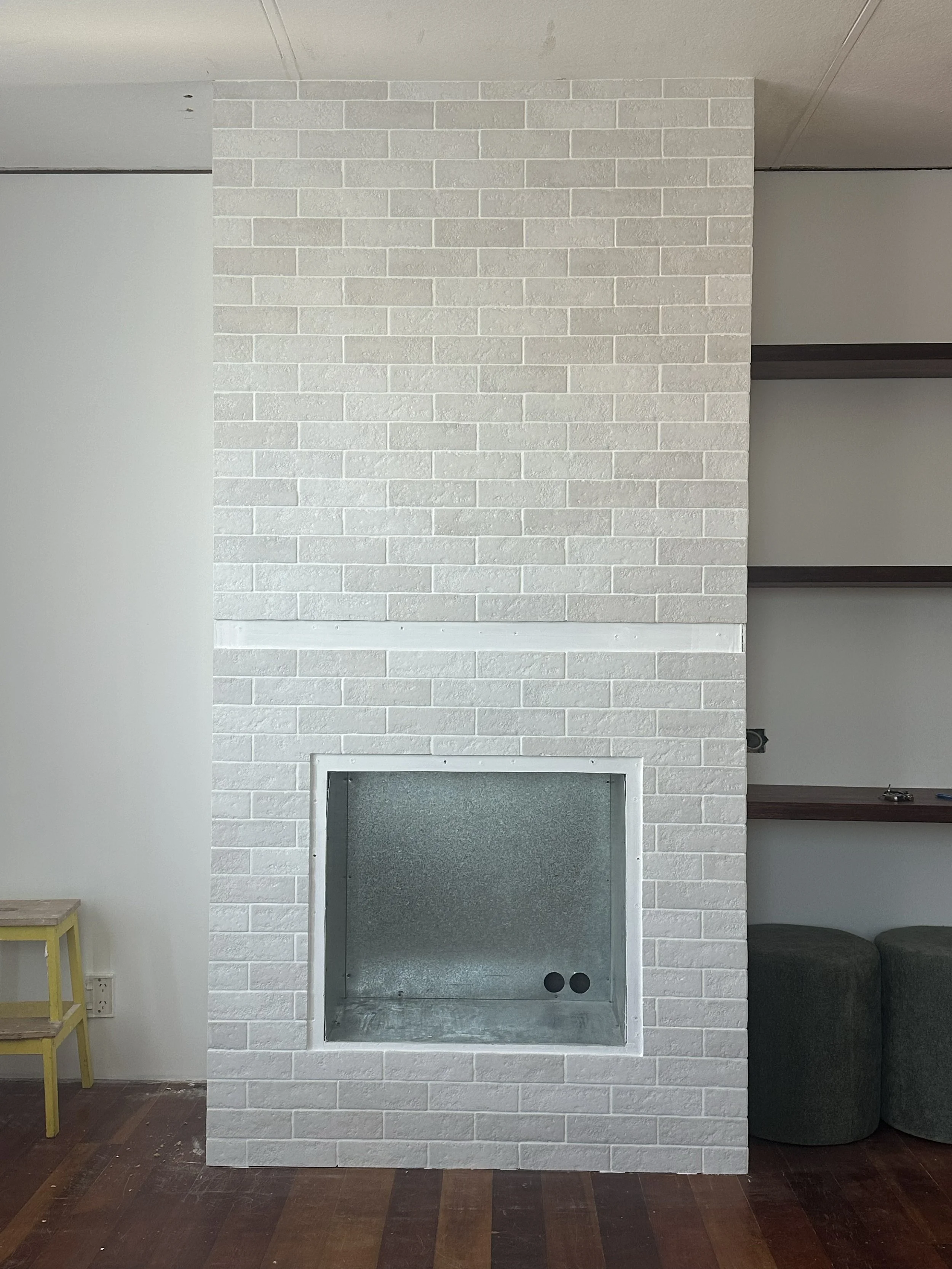 A white brick fireplace with a metal interior, situated in a room with wooden flooring and built-in shelves on the right side.