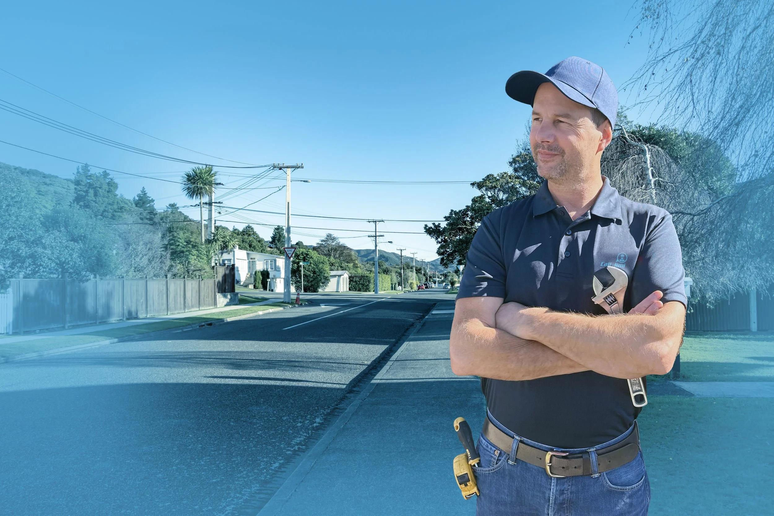 A man in a gray polo shirt and cap holding a wrench and a screwdriver, standing on a suburban street with crossed arms, sidewalk, fence, trees, and utility poles under a clear blue sky.