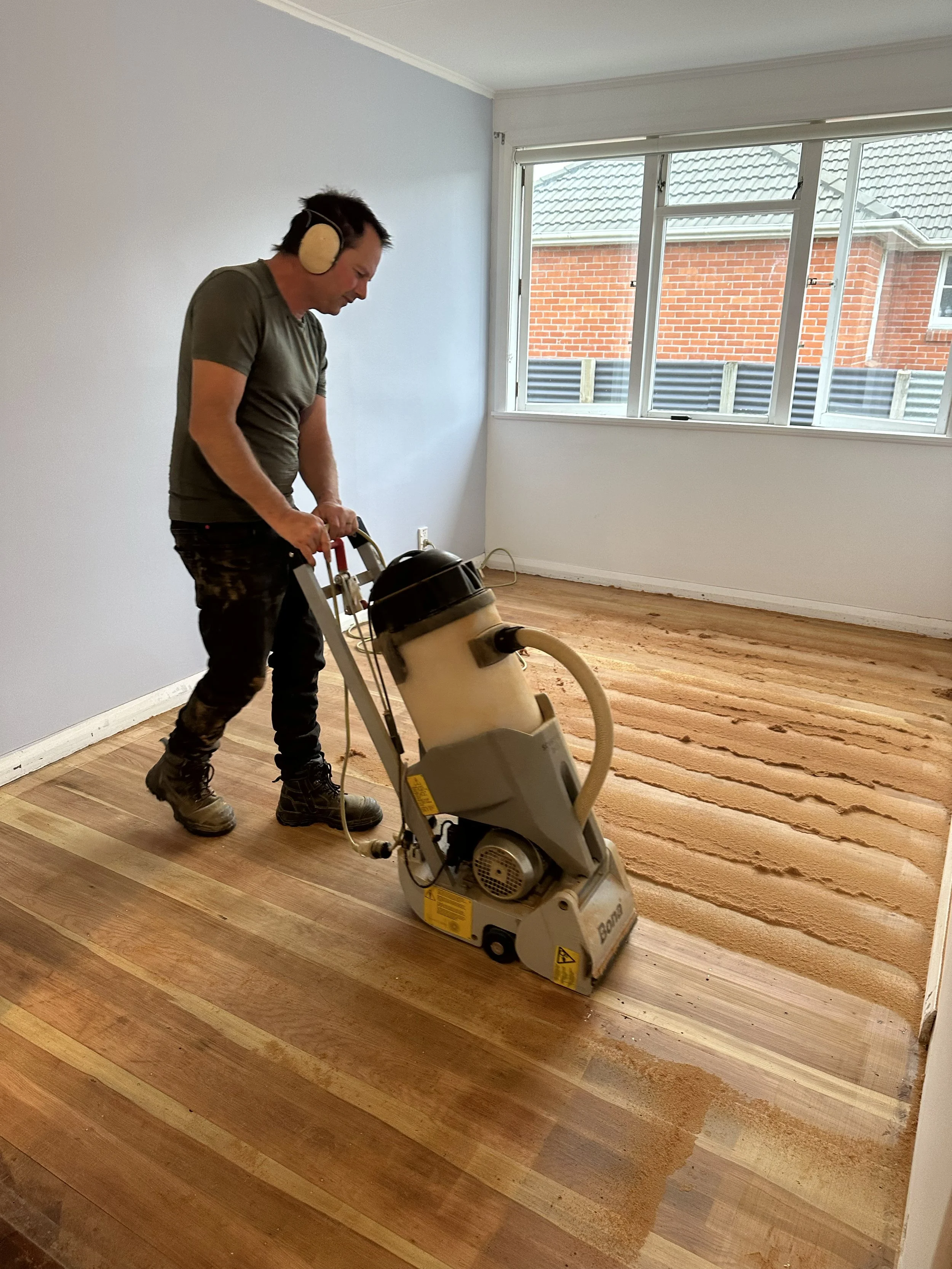 A man in protective headphones operating a floor sander on a hardwood floor in an empty room with white walls and a large window.