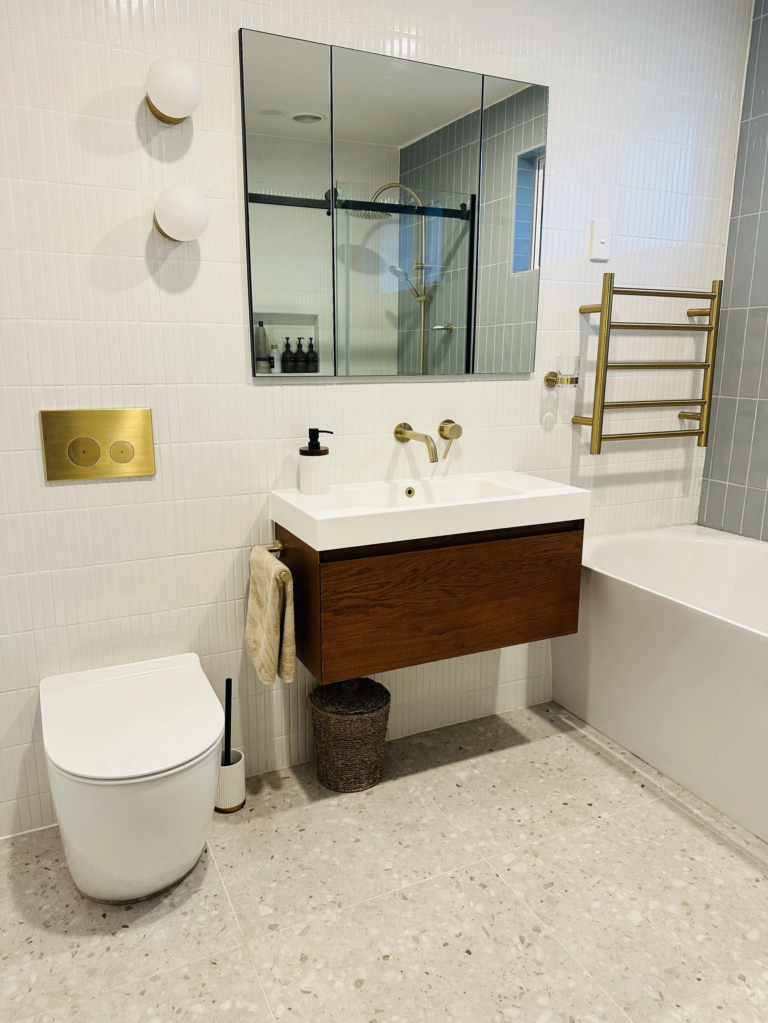Modern bathroom with white tiled walls, a wall-mounted sink with a wooden cabinet, a large mirror, a white toilet, a wicker wastebasket, a bathtub, a heated towel rack, and a walk-in shower with glass doors.