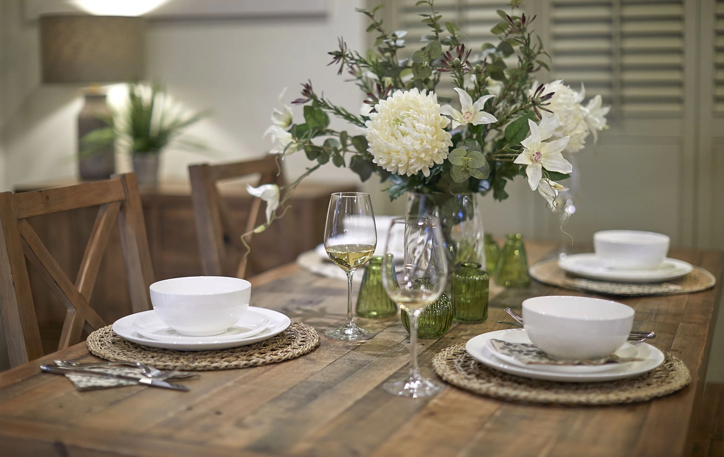 Dining table commercial editorial photograph with white crockery and a flower centre piece. Shallow depth of field with an out of focus lamp and sideboard behind the table.