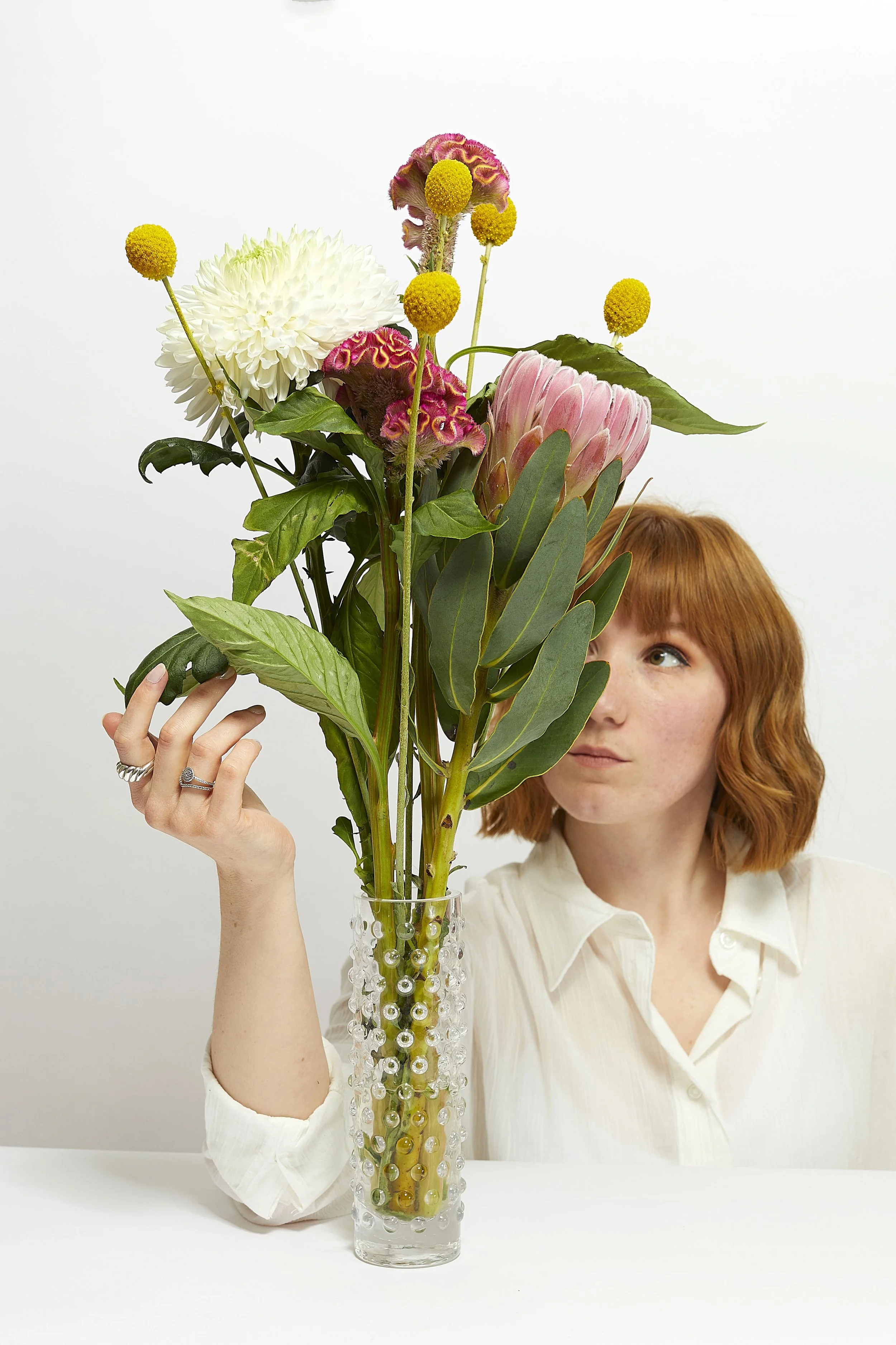 Self portrait studio photography, with a soft commercial aesthetic. Soft lighting, white background and white shirt styling editorial photo shoot. Posing with flowers, white pink and yellow colours.