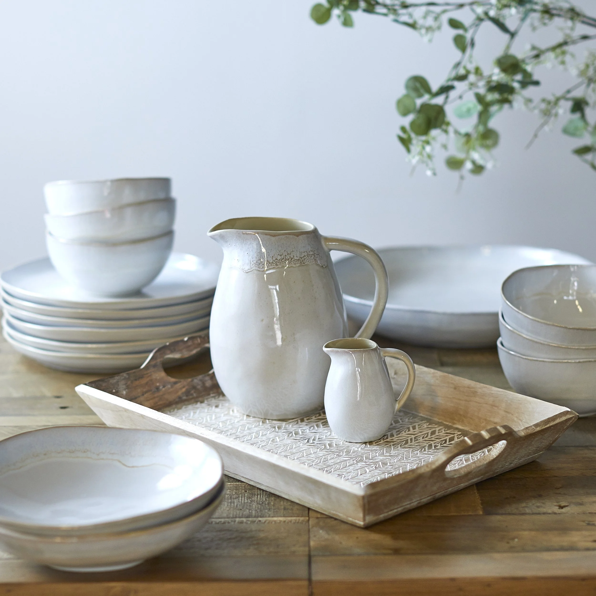 Commercial crockery still life photograph. Two jugs on a tray in the centre of the image with matching plates and bowls stacked around on a wooden table. Simple composition and shot with studio flash diffused to give bright highlights and soft shadow
