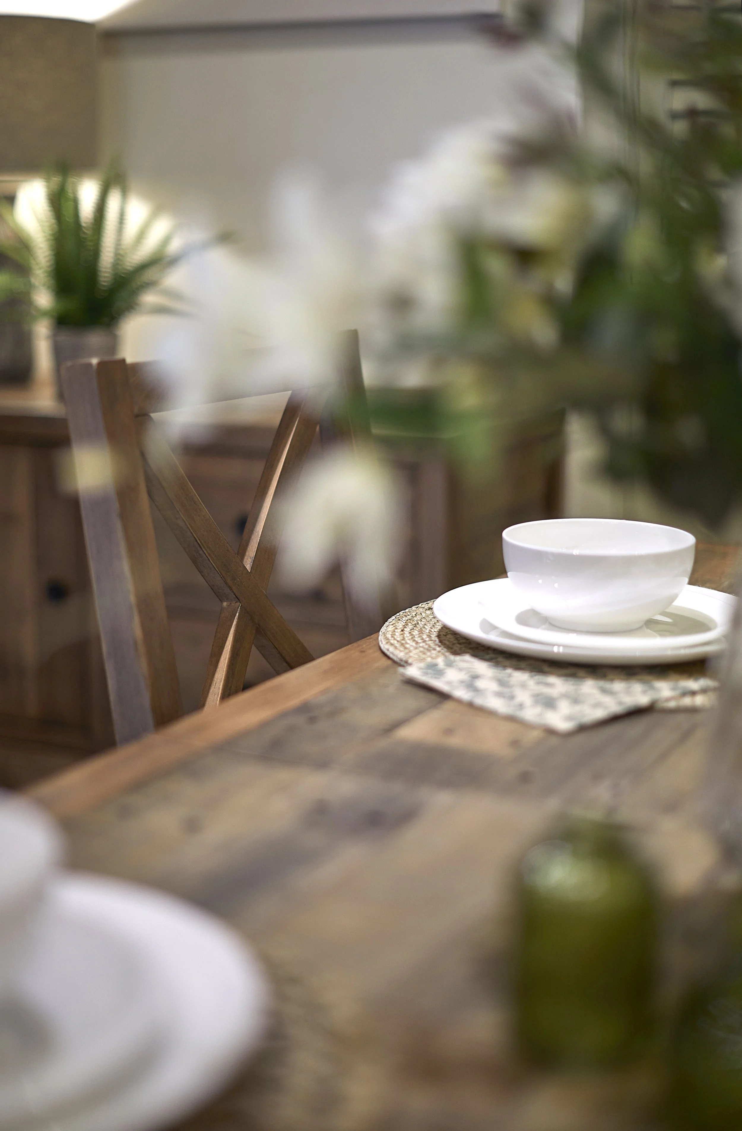 Dining table commercial editorial photograph with white crockery and a flower centre piece. Shallow depth of field with an out of focus lamp and sideboard behind the table.