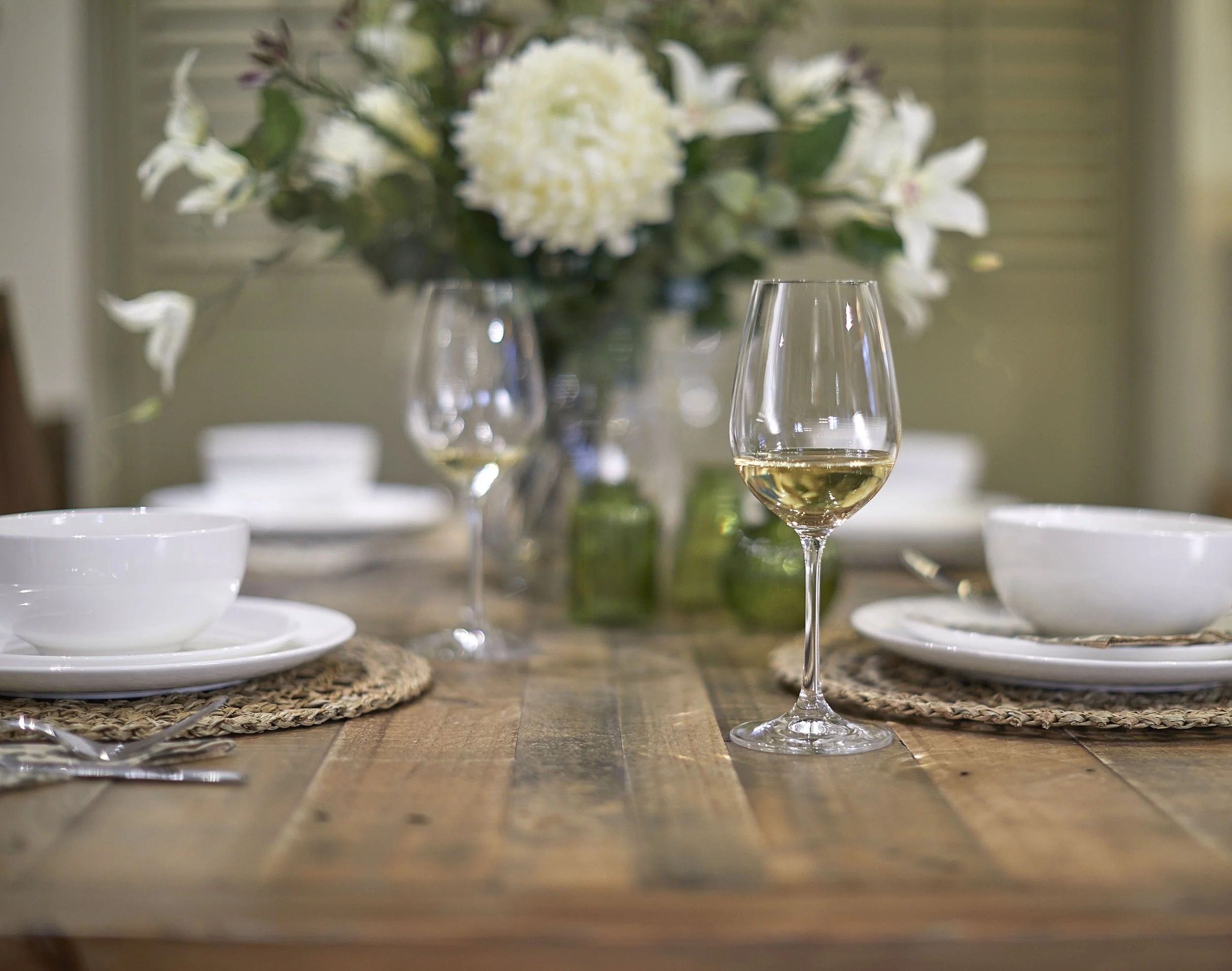 Dining table commercial editorial photograph with white crockery and a flower centre piece. Shallow depth of field with an out of focus lamp and sideboard behind the table.