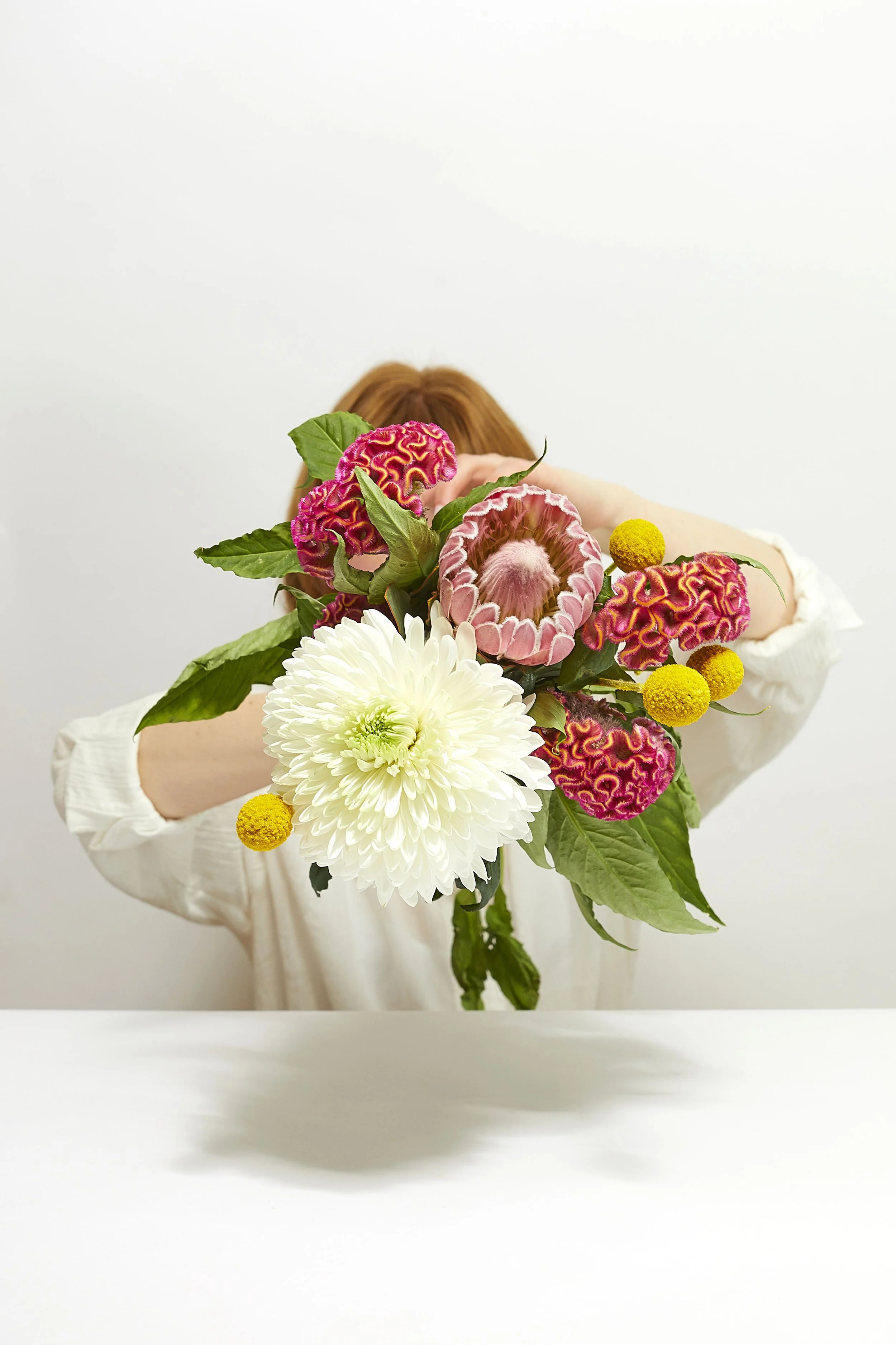 Self portrait studio photography, with a soft commercial aesthetic. Soft lighting, white background and white shirt styling editorial photo shoot. Posing with flowers, white pink and yellow colours.