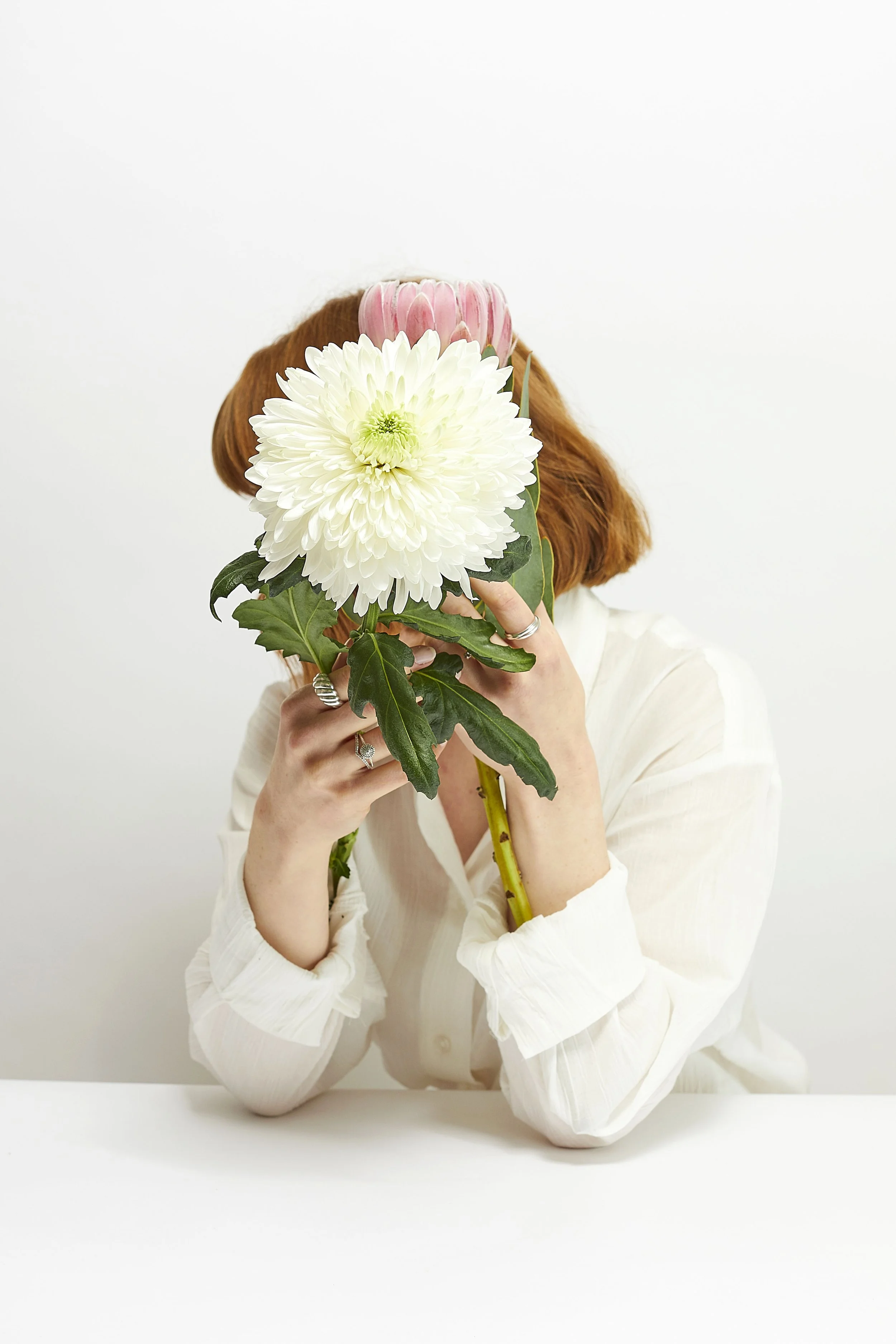 Studio self portrait photograph posing with flowers. Soft beauty editorial aesthetic, bright lighting with white backdrop, focus on flowers and soft focus on face