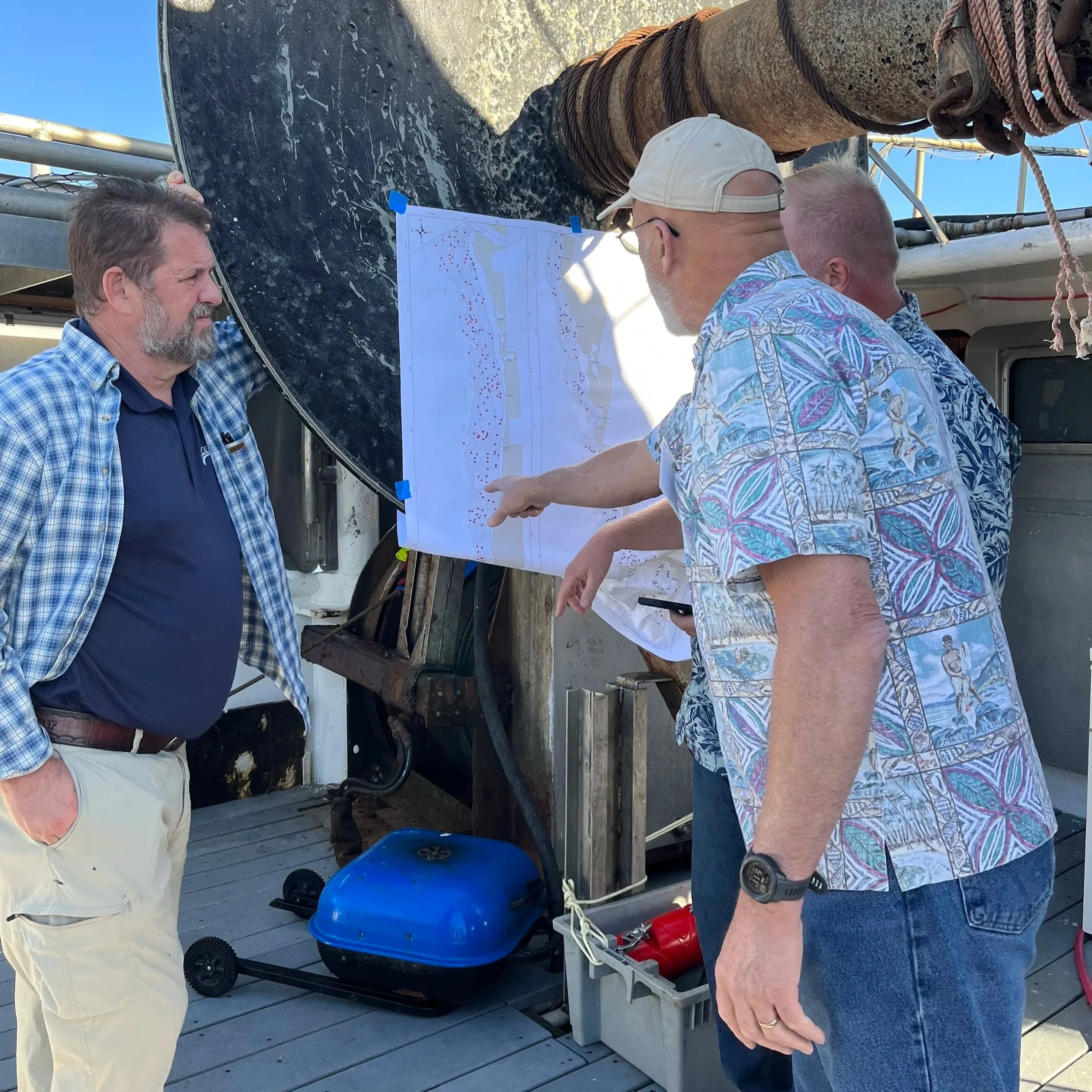 Three men standing outdoors near a large satellite dish. One man in a checked shirt is looking at a diagram posted on the dish, while the other two men, one wearing a patterned shirt and cap, are pointing at the diagram and discussing. The scene appe