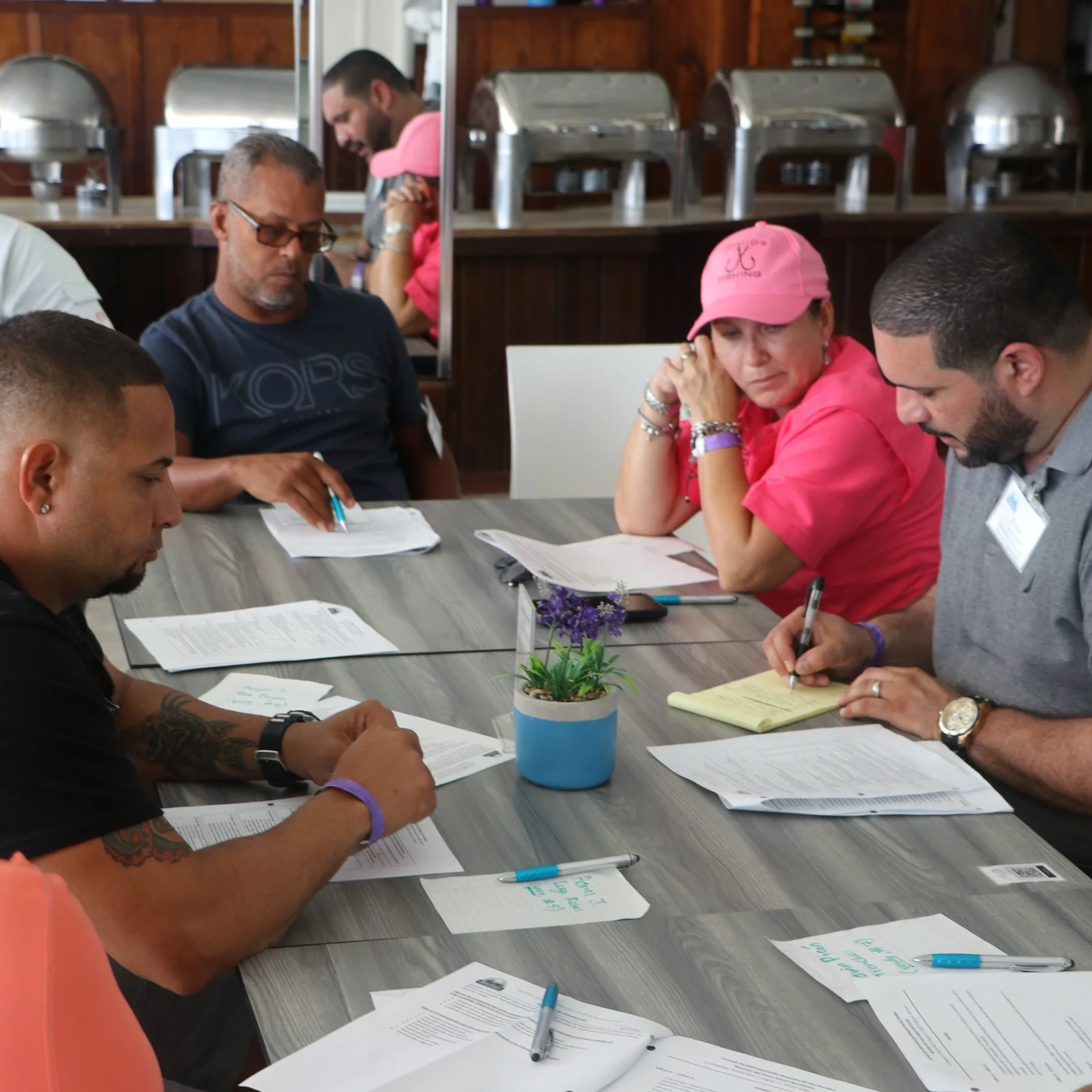Group of people sitting around a table in a meeting, reviewing documents, with a potted plant in the center of the table.