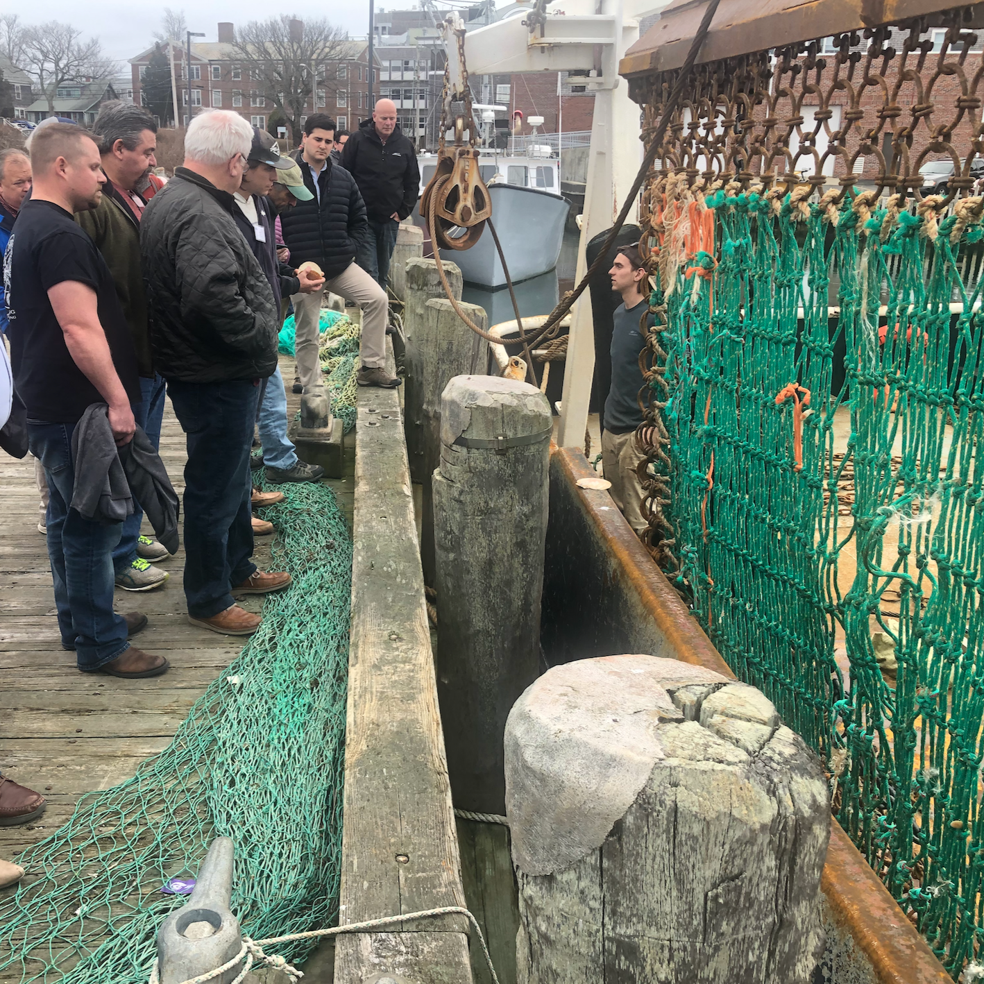 A group of people standing on a wooden dock, observing a man who is talking to them. The dock has green fishing nets along the edge, and there are rocks, a boat, and nautical equipment nearby. The background shows houses and trees.