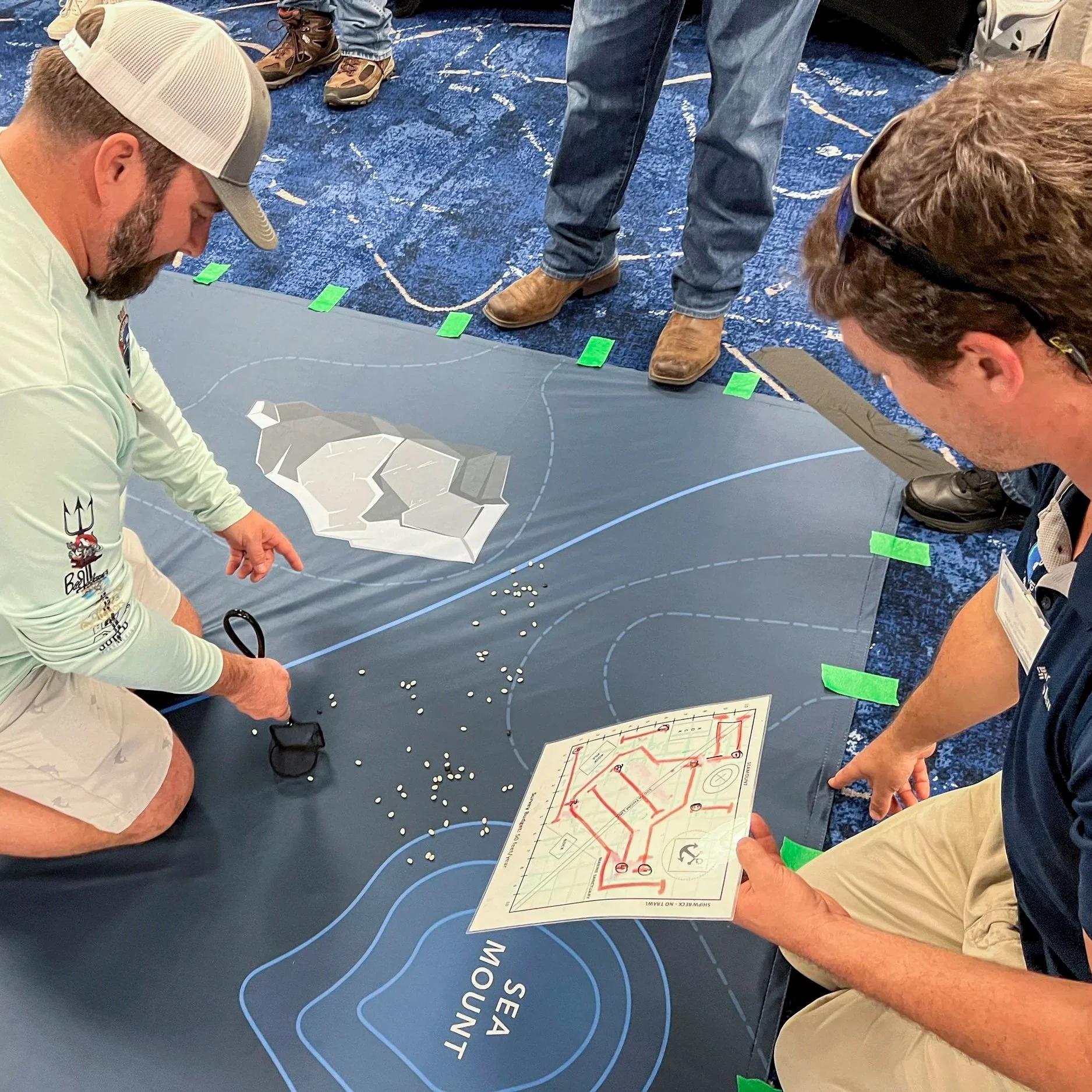 Two men kneel on a large blue map of the Sea of Maine, pointing at small white balls, possibly representing fish or objects. One man holds a laminated map of a baseball field. Several people stand around observing.